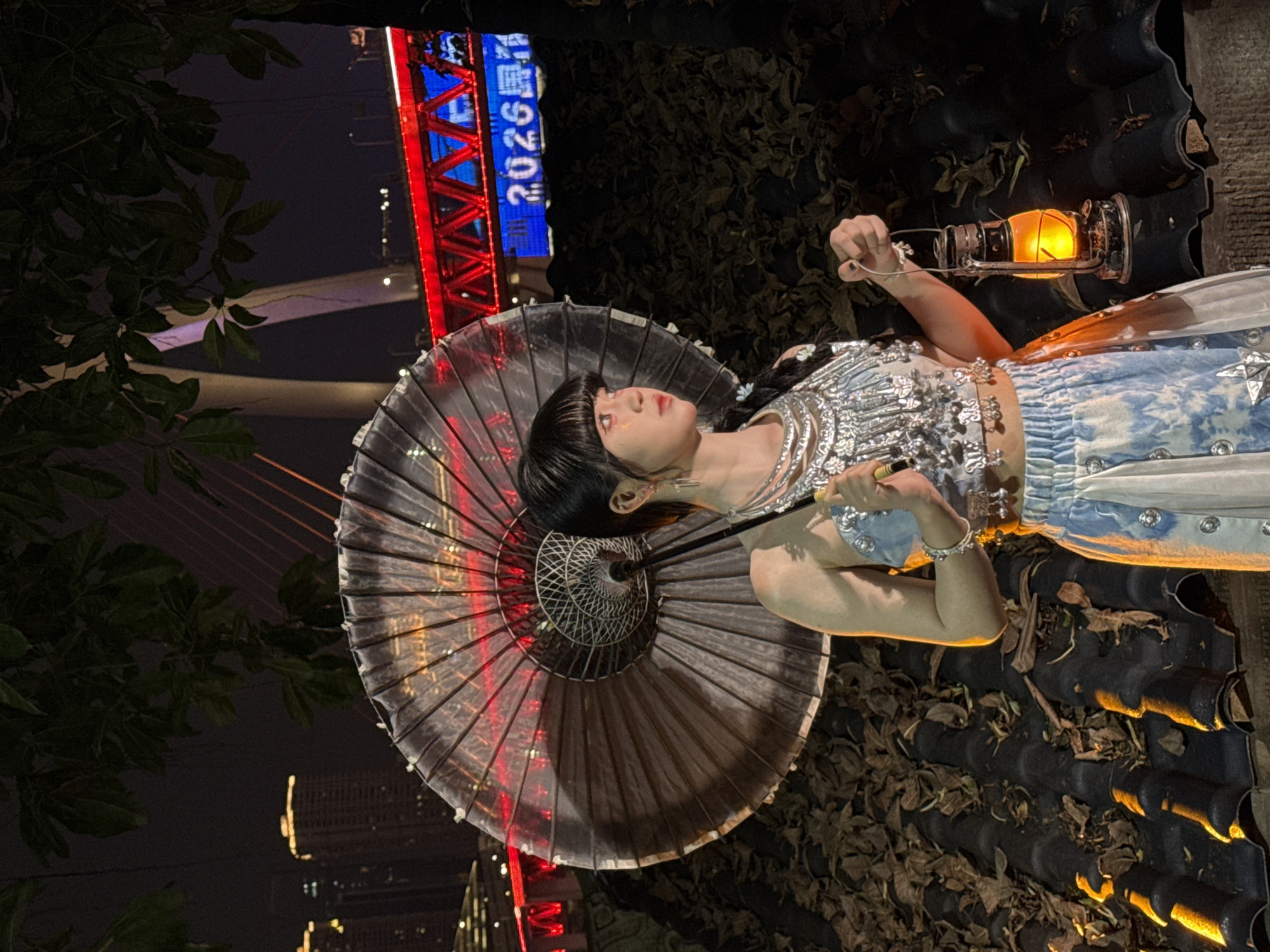 A young woman with black hair and bangs is holding a gray umbrella with red lights while looking up to the left. She is wearing a silver top, a blue and white skirt, and jewelry on her wrists and ears. In her right hand, she holds a lit lantern. The background features a cityscape at night, with a large bridge illuminated in red and blue. The blue lights display the numbers "2026." The woman is positioned on a surface of dark tiles with scattered leaves. The image is taken at night in the Yuzhong District of Chongqing, China.