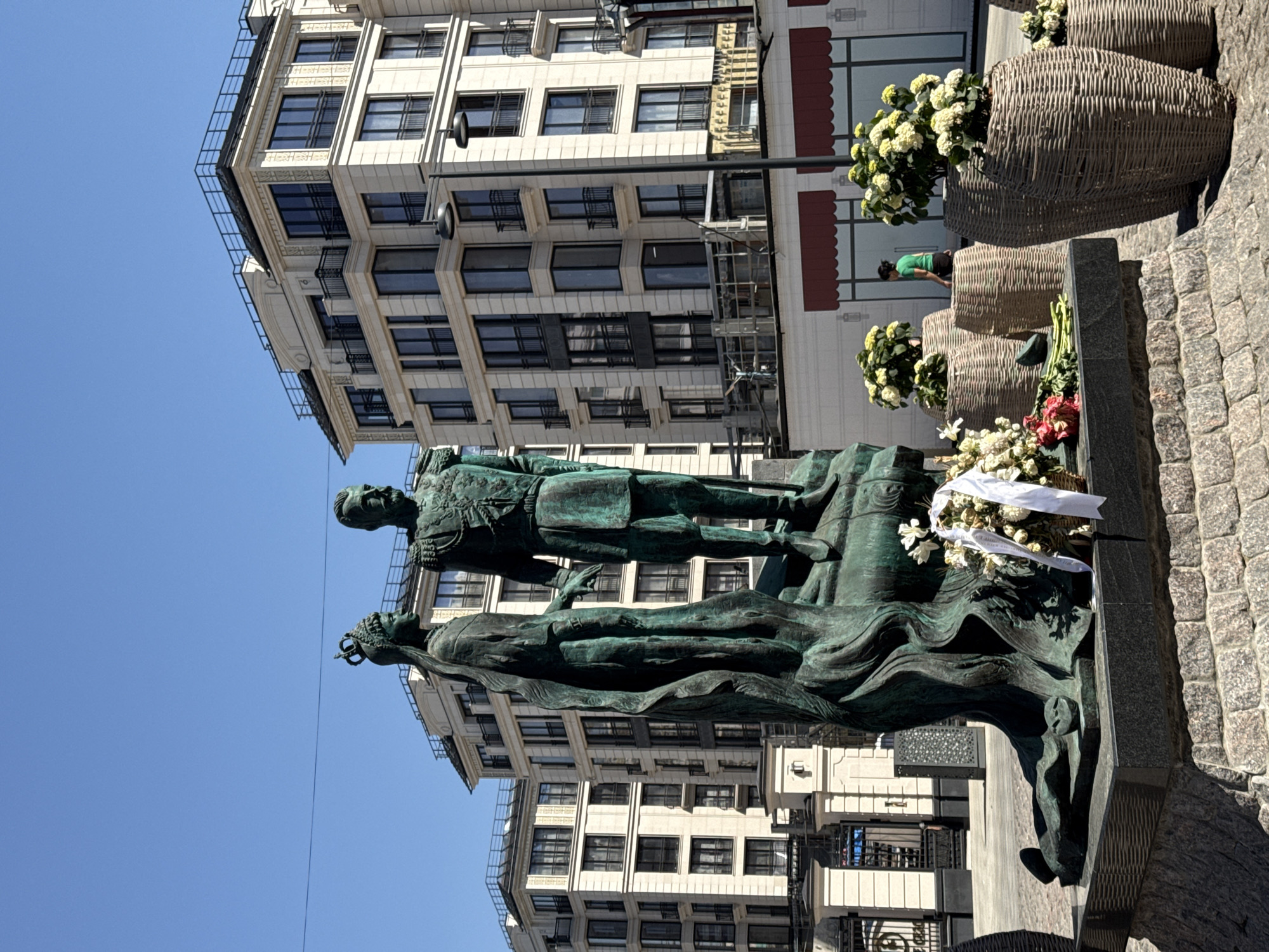 The image shows a bronze statue of a man standing next to a woman lying down on a stone pedestal. The man is wearing a military uniform, and the woman is wearing a crown. A building with many windows is in the background. There are also large flower baskets next to the statue. The statue is likely located in a city square or park. There are flowers placed on the statue.