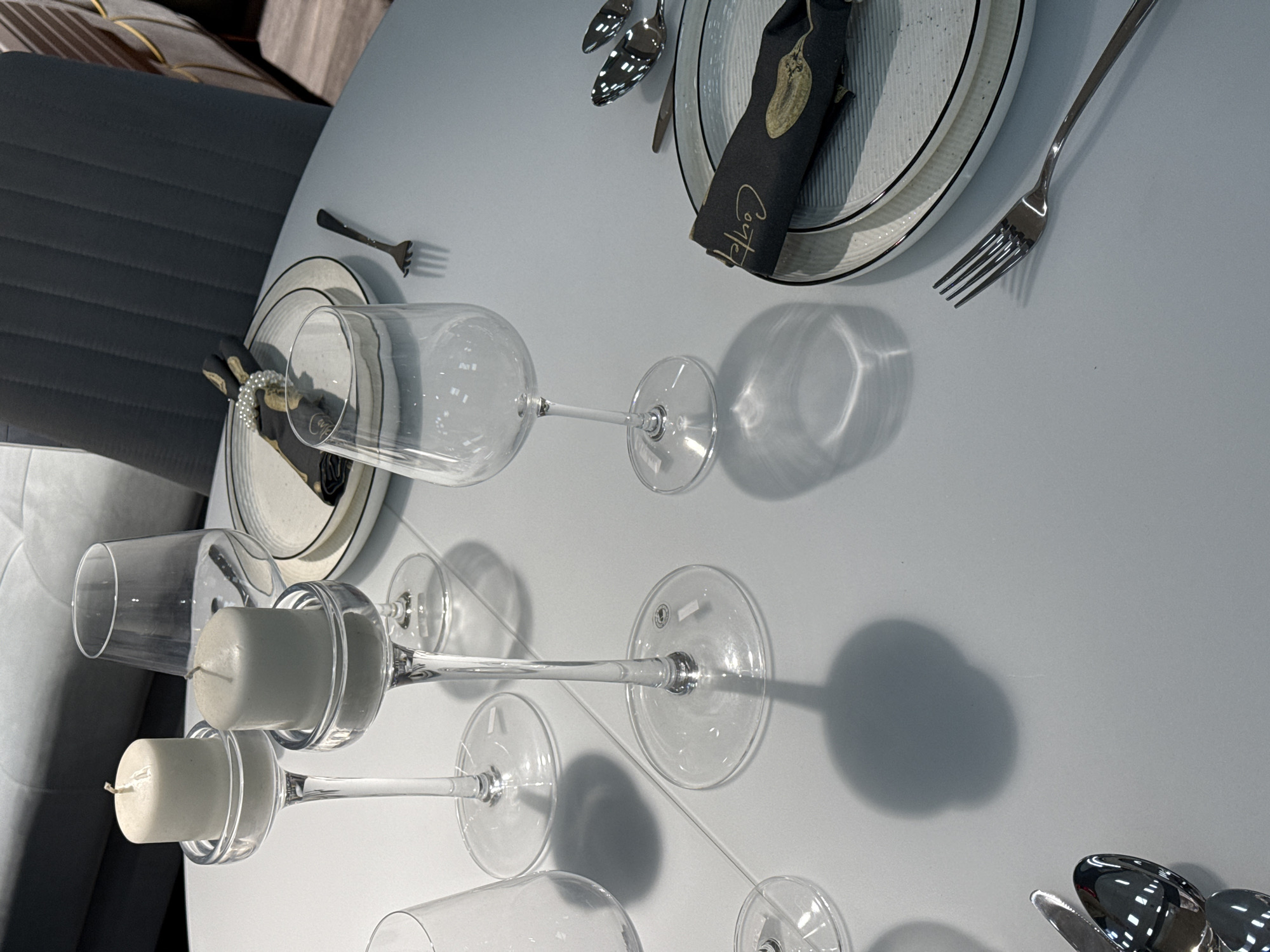 The scene captures a setting on a round, light grey table. There are dinnerware elements including plates, cutlery (knives and forks), and glasses, some with candles set within. The plates are predominantly white with black rims, and a black napkin with gold lettering is placed on one of the plates. Silver cutlery is arranged around the plates. A few clear wine glasses and candlestick holders are present, some holding white candles. The lighting is ambient. The location is indicated as Izhevsk, Russia.
