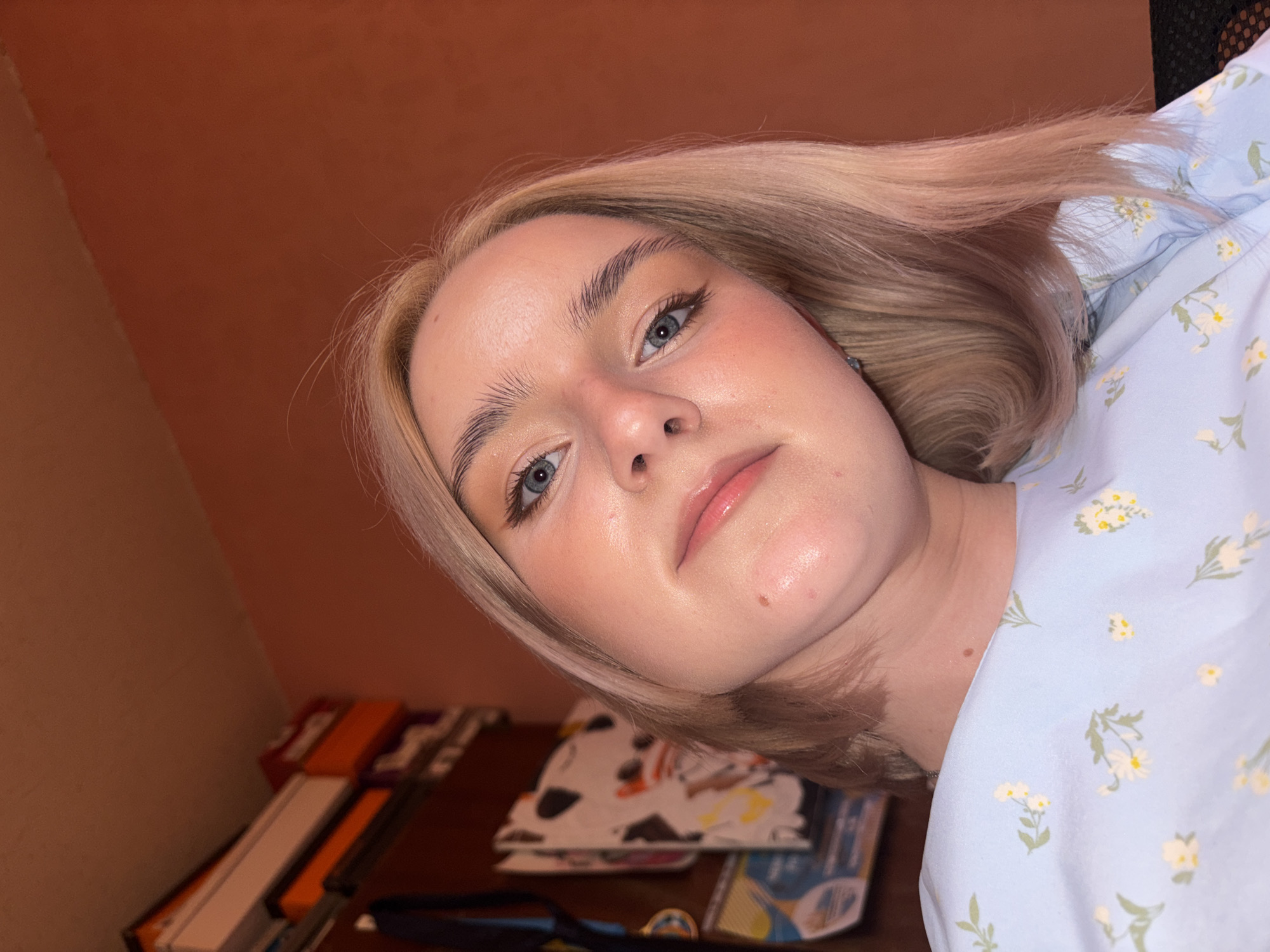 A young female is captured in a close-up, upward-angled selfie. She has light, possibly ash blonde, hair styled with some volume, and striking blue eyes. Her complexion is fair, enhanced with visible blush on her cheeks, a subtle lip gloss, and defined eyebrows with light eyeliner. Her head is tilted slightly to her right, and she looks directly at the camera with a calm, neutral expression.
She is dressed in a light blue top or dress patterned with small white and yellow daisy-like flowers and green foliage. The background behind her consists of a solid, warm reddish-brown wall. Below her, a wooden desk or surface is visible, partially obscured and out of focus. On this surface, various objects are stacked, including books or binders with prominent orange spines, alongside a white item featuring black and orange designs. The lighting in the indoor setting is warm, illuminating her features and hair. No text is visible in the image.