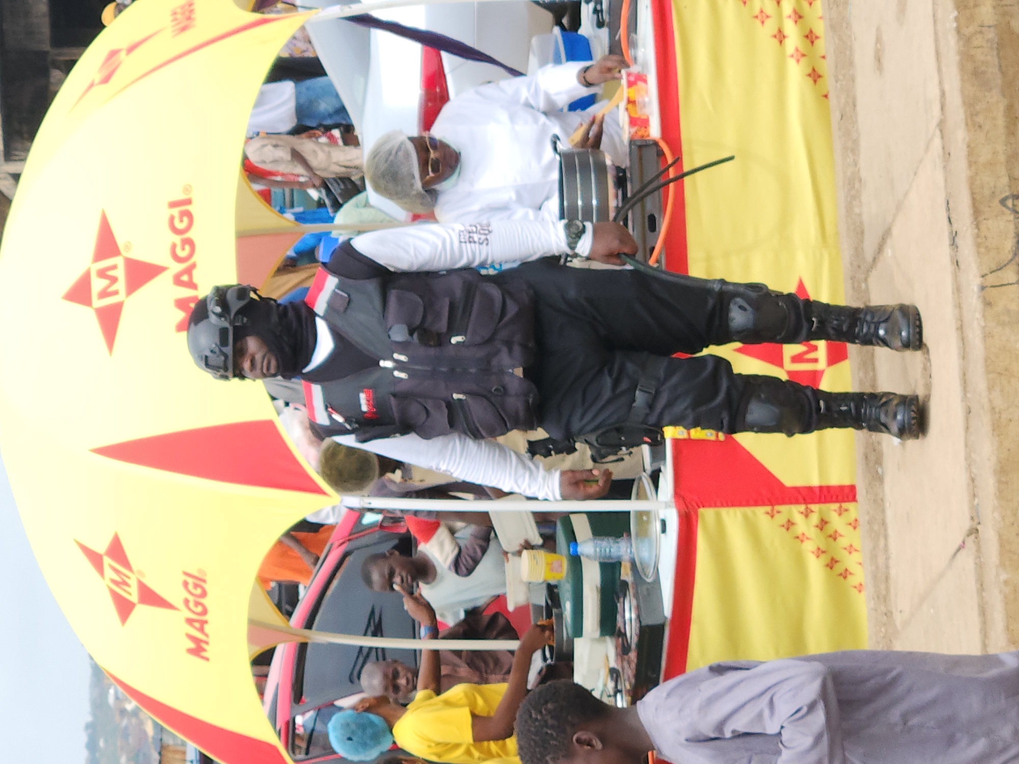 The image shows a man in a black tactical vest, helmet, and boots standing in front of a yellow and red Maggi-branded structure. He is wearing a white shirt underneath the vest. Inside the structure, a man in a white shirt and hairnet is working, while other individuals are seated or standing around. The location is Jos, Nigeria.
