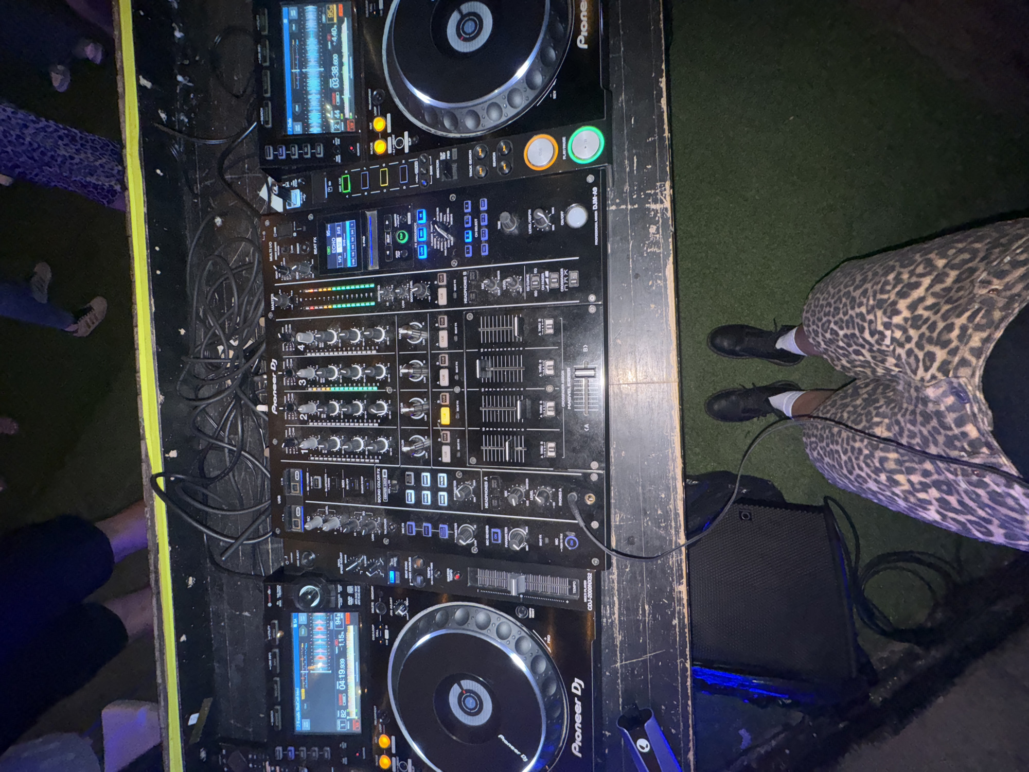 A high-angle, eye-level shot captures a DJ setup, likely in an outdoor, grassy area. The DJ booth is constructed from dark wood, with a yellow band across the top. The central focus is a Pioneer DJ setup, featuring two CDJ-3000 media players and a DJM-A9 mixer. The media players display waveform data, track times, and bpm. Various knobs, buttons, and sliders are visible on the mixer, indicating active use. Cables are neatly organized on top of the booth.
In the foreground, the lower body of a person is visible, wearing leopard-print pants and dark shoes with white socks. This individual is positioned behind the DJ equipment, suggesting they are the DJ. In the background, partially obscured, are the legs of other individuals, indicating an event or gathering is taking place. The ground is covered in artificial turf, resembling grass. No specific Moscow landmarks or street signs are present, making precise location identification impossible based on visual cues alone. The overall scene suggests a performance or party setting.