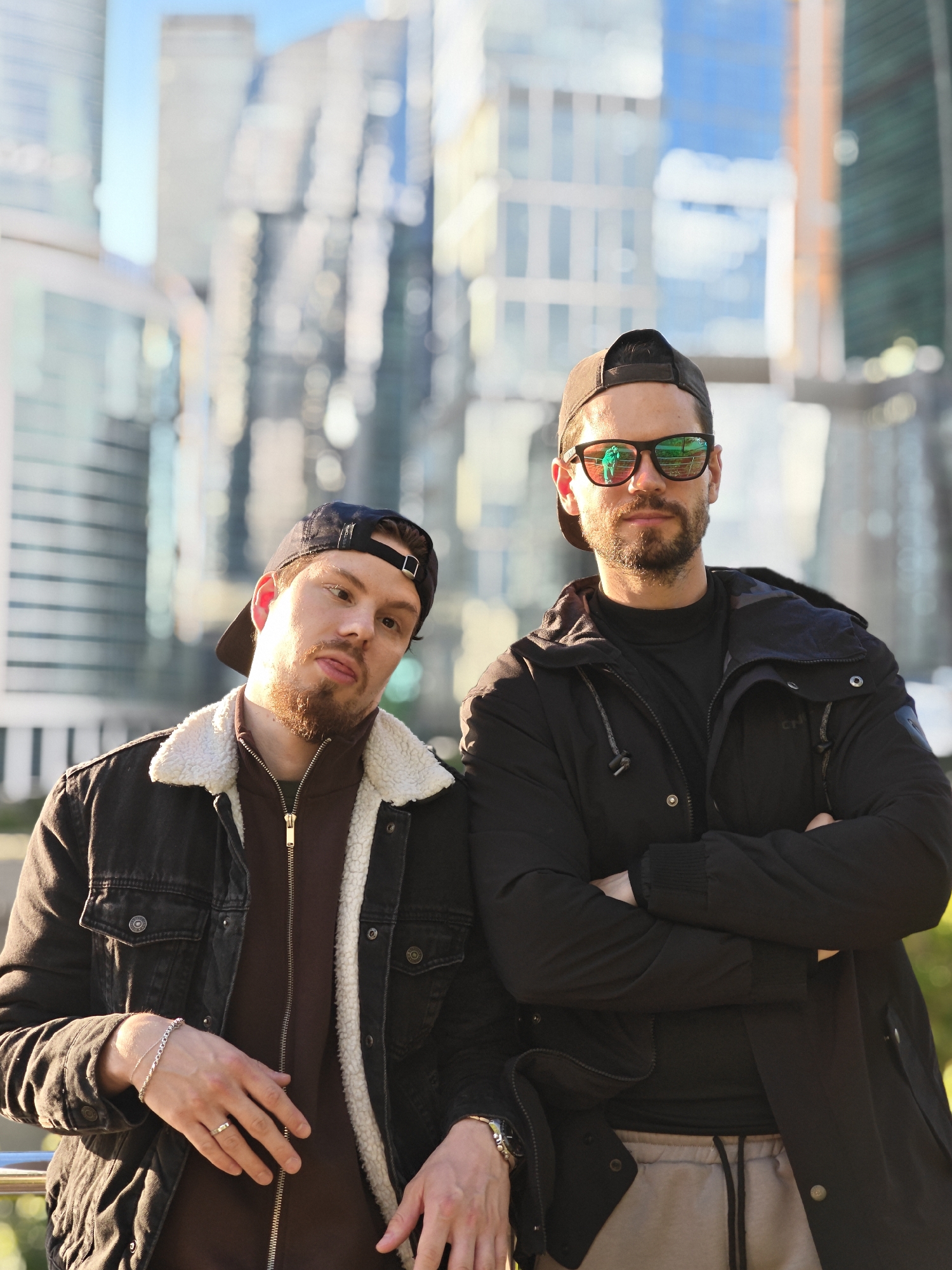 Two men are standing close together in an outdoor setting with a backdrop of modern high-rise buildings. The buildings suggest an urban environment, likely a business district. The image appears to be taken in the city of Moscow, Russia, based on the architectural style of the buildings.
The man on the left has a beard and is wearing a black baseball cap and a dark denim jacket with a light-colored, fleece-lined collar. He has his left hand resting on the railing. He wears a wedding ring and a bracelet.
The man on the right is wearing sunglasses, a black baseball cap, a black jacket, and a black shirt. He has his arms crossed.
The lighting suggests it is daytime. The details of the background buildings are somewhat blurred, indicating a shallow depth of field.