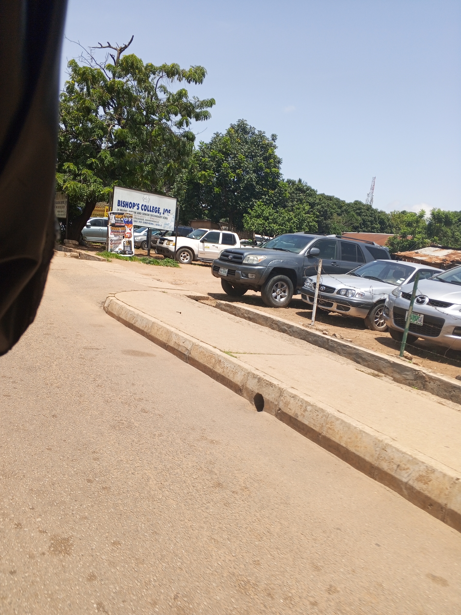 The image shows a street scene in front of Bishop's College, Jos, Nigeria. The foreground is dominated by a dusty road with a concrete curb. Beyond the curb, several vehicles are parked on a dirt lot. These include a dark gray SUV, a silver sedan, and a white pickup truck. A sign for Bishop's College, Jos, is visible behind the parked cars. The background features lush green trees and a clear blue sky, with a distant cell tower. The overall impression is a sunny day in a semi-urban or suburban setting.