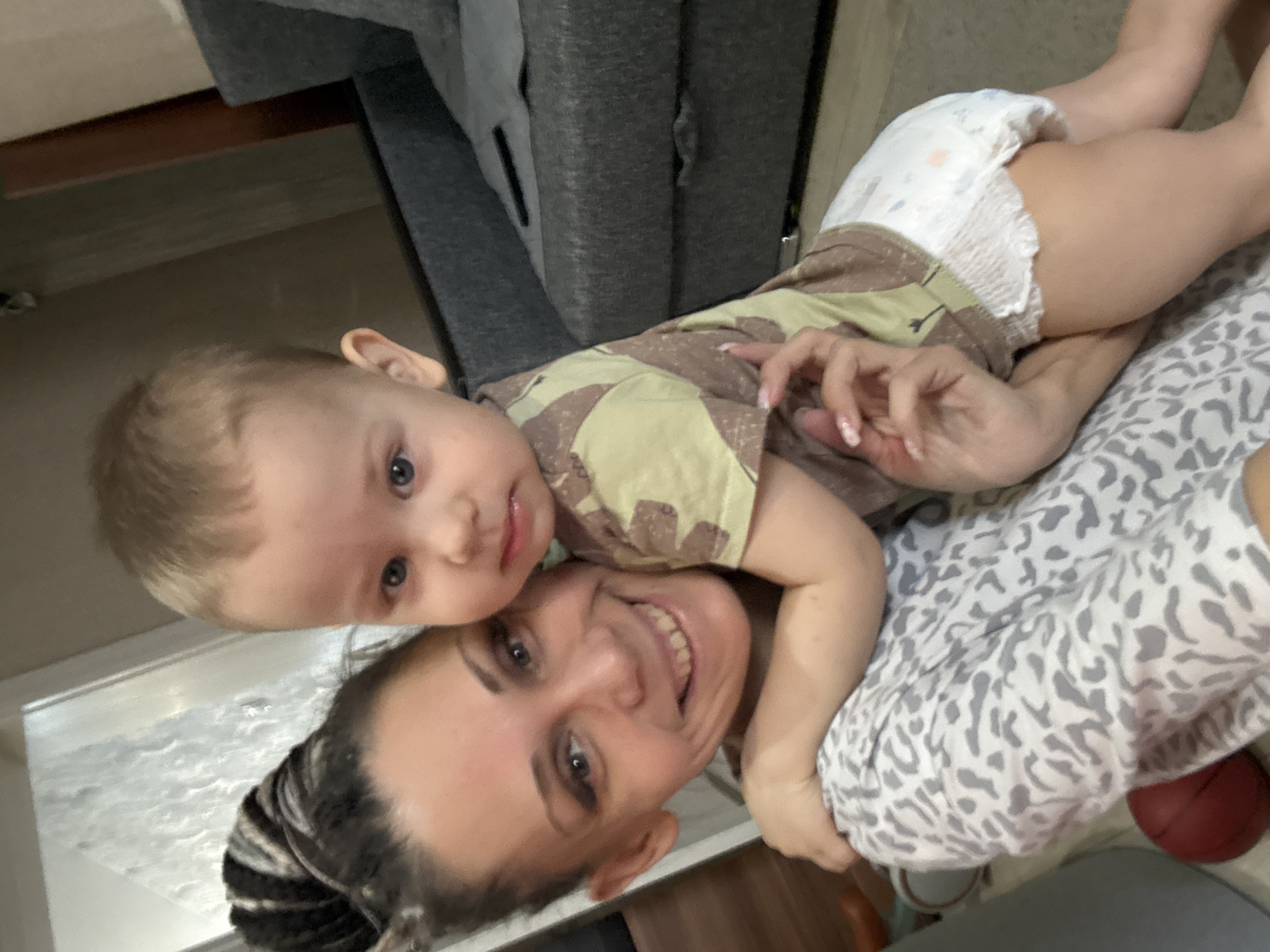 The image shows a woman and a baby. The woman is smiling and has dark hair pulled back. The baby is wearing a camouflage shirt and a diaper, and is lying on top of the woman. The background is out of focus, but appears to be indoors. A red ball can be seen under the clothes the woman is wearing. The location appears to be Unknown, Unknown.