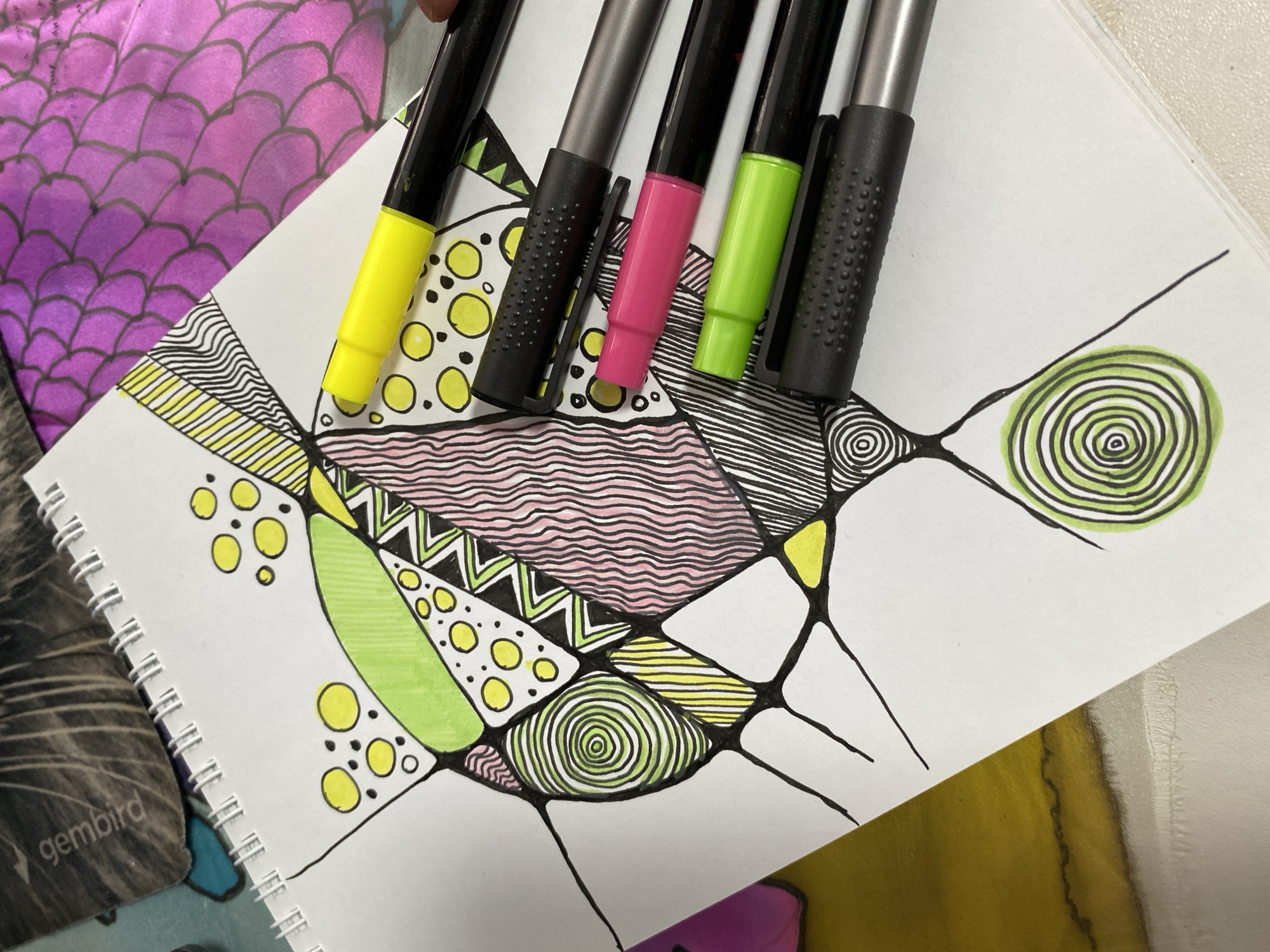 The image displays an artistic scene featuring a vibrant, abstract doodle drawing and a collection of markers, set in Noviy, Russia. The central subject is a geometric drawing on white paper within a spiral-bound sketchbook. Bold black lines divide the composition into irregular shapes, which are filled with diverse patterns, including parallel lines, wavy stripes, concentric circles, and scattered dots. The dominant colors within the artwork are bright yellow, lime green, and a soft pink, creating a lively contrast against the black outlines.
Five markers are positioned across the drawing, suggesting their use in its creation. They feature black bodies with distinct colored caps or tips: a bright yellow, a hot pink, a lime green, and two with textured black grips (one with a black tip and another with a silver/grey tip).
In the upper left corner, a portion of a fabric with a purple and magenta gradient and a distinctive scalloped or scale-like pattern is visible, providing a contrasting texture and color. The lower-left corner reveals the binding and part of the black cover of the spiral-bound sketchbook, where the brand name "gembird" is subtly visible. The scene captures a moment of creative activity, focusing on the intricate details of the art and the tools used. The lighting indicates an indoor setting.