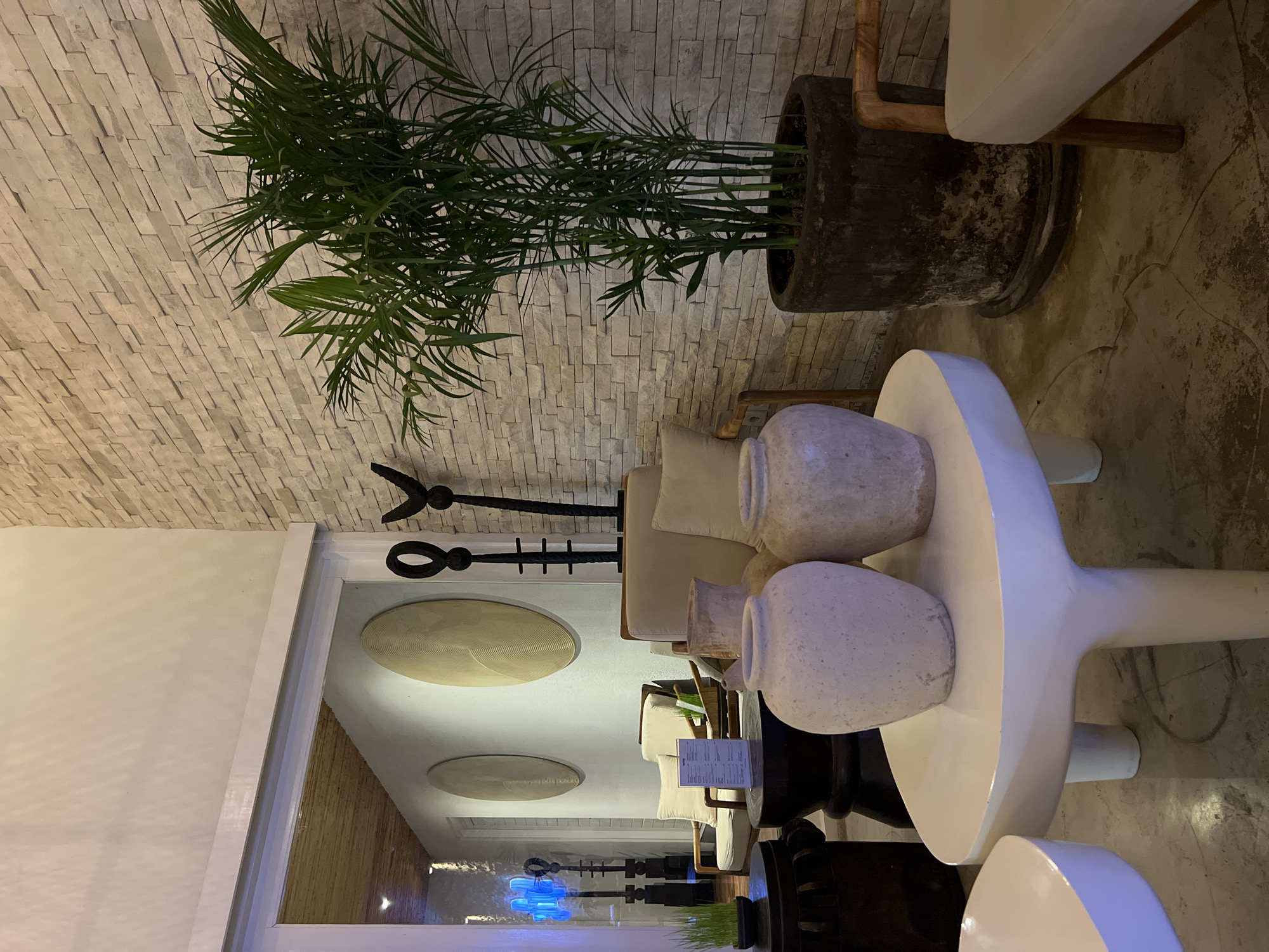 The image shows an interior space with a plant in a large pot in the upper right corner and a stone wall behind it. A white table with multiple decorative urns sits near the center. An elongated doorway reveals a sitting area with chairs and decorative wall pieces. In the distance, an illuminated art piece is visible. Based on the tags, this location is in North Kuta, Indonesia.