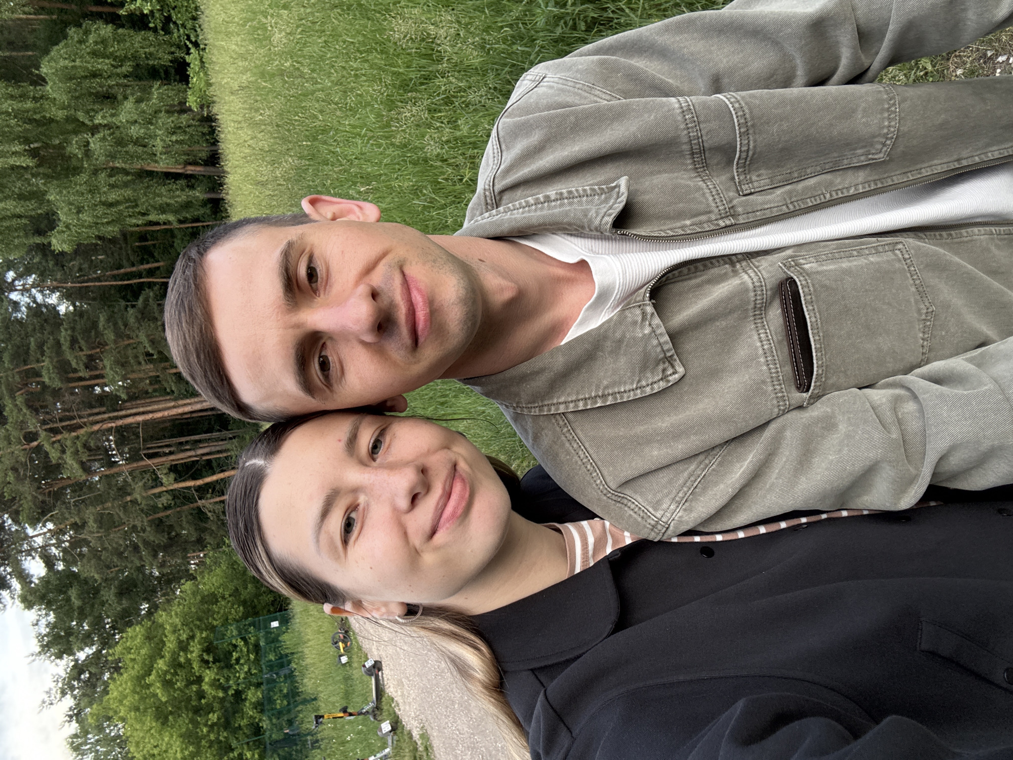 An outdoor medium shot depicts a young Caucasian couple posing for a selfie in a park. The woman is positioned on the left, facing the camera with a slight smile. She has blonde hair pulled back and is wearing a black jacket over a striped shirt. The man is on the right, also facing the camera with a relaxed expression. He has short brown hair and is wearing a green jacket over a white t-shirt. They are standing close together, suggesting an intimate relationship.
The background reveals a park environment with a gravel path on the left, where several electric scooters are parked. Tall green trees form a dense forest behind the couple, and a field of tall green grass is visible to the right. The lighting suggests it is daytime, possibly late afternoon, given the soft, natural light. The overall impression is a casual and pleasant outdoor outing. This image does not provide any specific details to locate it within Moscow, Russia.