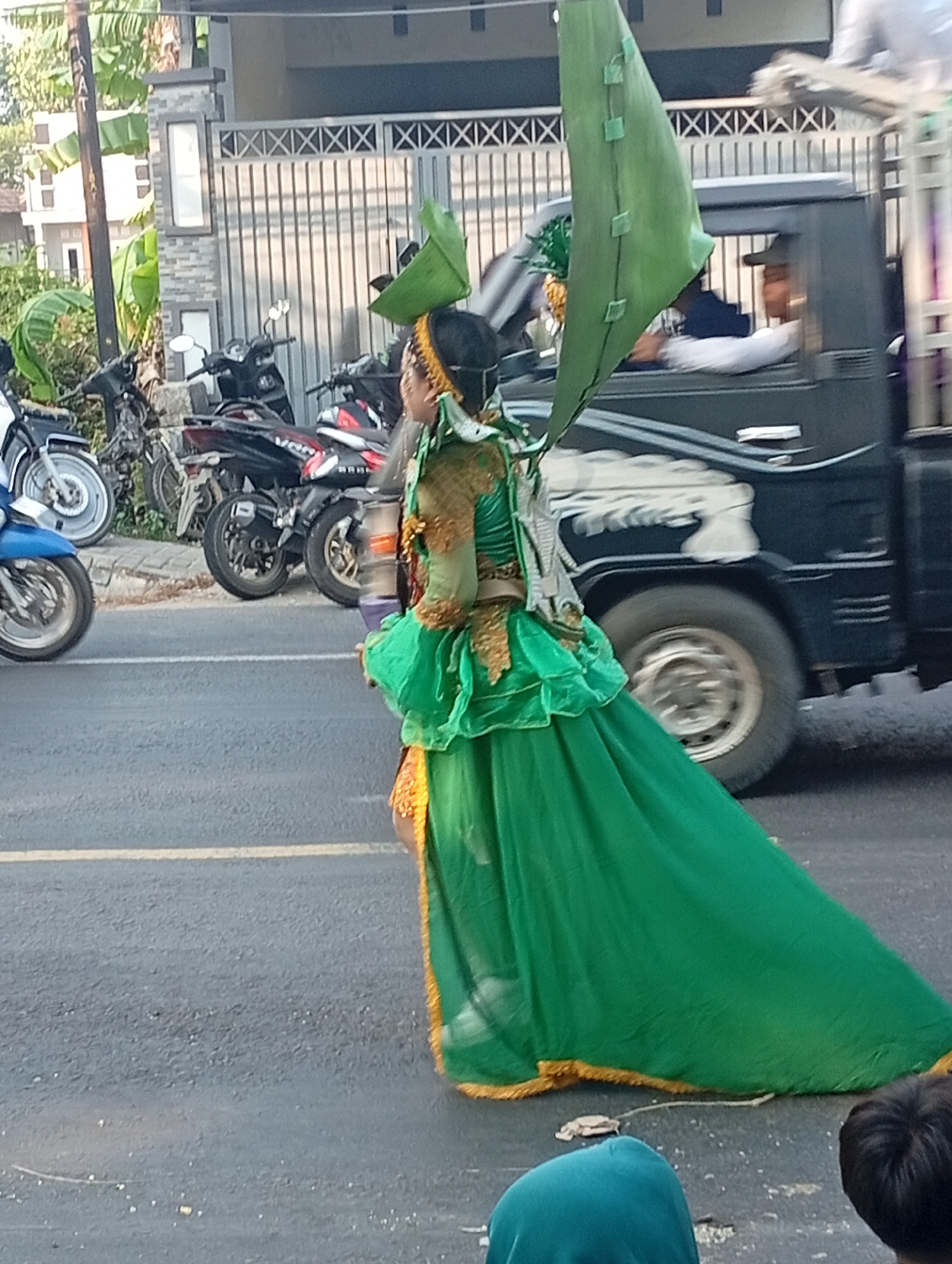 In the city of Bajingjowo, Indonesia, a person adorned in a vibrant green costume with gold accents walks along a paved street. The costume features a flowing skirt and elaborate upper body detailing, topped with a large, leaf-like green headpiece that extends over their shoulder. Behind the person, several motorcycles are parked along the side of the road. A dark-colored truck with a white graphic on its side is visible in the background, and a person wearing a baseball cap can be seen through the truck's window. The scene appears to be part of a parade or festival, given the festive attire and the presence of onlookers, one of whom is partially visible at the bottom right of the frame. The environment suggests a warm, sunny day, with a building and vegetation visible in the far background.
