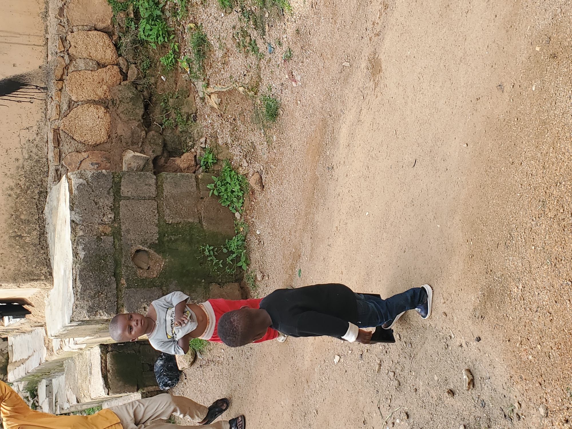 Two young boys are standing in a dirt area next to a stone and brick wall. The boy on the left is wearing a light gray t-shirt and red shorts, and he is holding up the hem of his t-shirt. The boy on the right is wearing a black long-sleeved shirt and dark blue jeans. He is holding a dark rectangular object in his left hand. Another person, partially visible at the bottom of the frame, is wearing light brown pants and a yellow top and appears to be wearing sandals. The ground is composed of sand and small pebbles, with some sparse green vegetation growing near the wall. The wall itself is constructed of rough-hewn stones and lighter-colored bricks, some of which appear worn and mossy. The scene suggests an outdoor setting in a somewhat rustic environment. There is no visible text. The lighting suggests it is daytime.