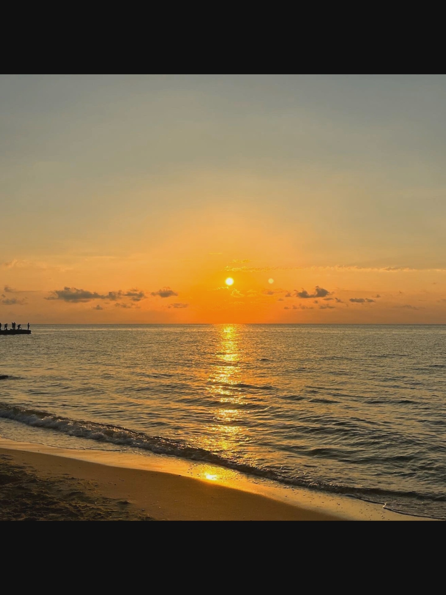 This is a photograph of a sunset over the sea, taken at the beach in Bielawska Wieś, Poland. The sun is setting on the right side of the frame, casting a golden glow across the water and the sky. There are several people silhouetted on a pier in the distance on the left. The waves are gently lapping at the sandy shore in the foreground. The sky is a gradient of orange and yellow, with a few scattered clouds. The water is a deep blue, reflecting the colors of the sunset.