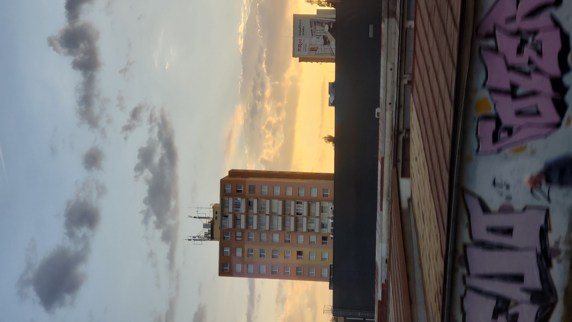 The image depicts a sunset scene in Poprad, Slovakia, with a tall, orange residential building dominating the center. The building has numerous windows and balconies and is topped with telecommunication antennae. To the right of the residential building, a smaller structure displays a billboard for "MiRad kúpeľnové štúdio" (MiRad Bathroom Studio). In the foreground, a dark, industrial-looking wall with graffiti runs horizontally across the lower portion of the frame. Above this, several rows of corrugated metal roofs of garages or storage units are visible. The sky is a mix of blue and orange hues, with scattered clouds illuminated by the setting sun. A person is partially visible walking in front of the graffiti-covered wall. The overall impression is one of an urban environment during twilight.