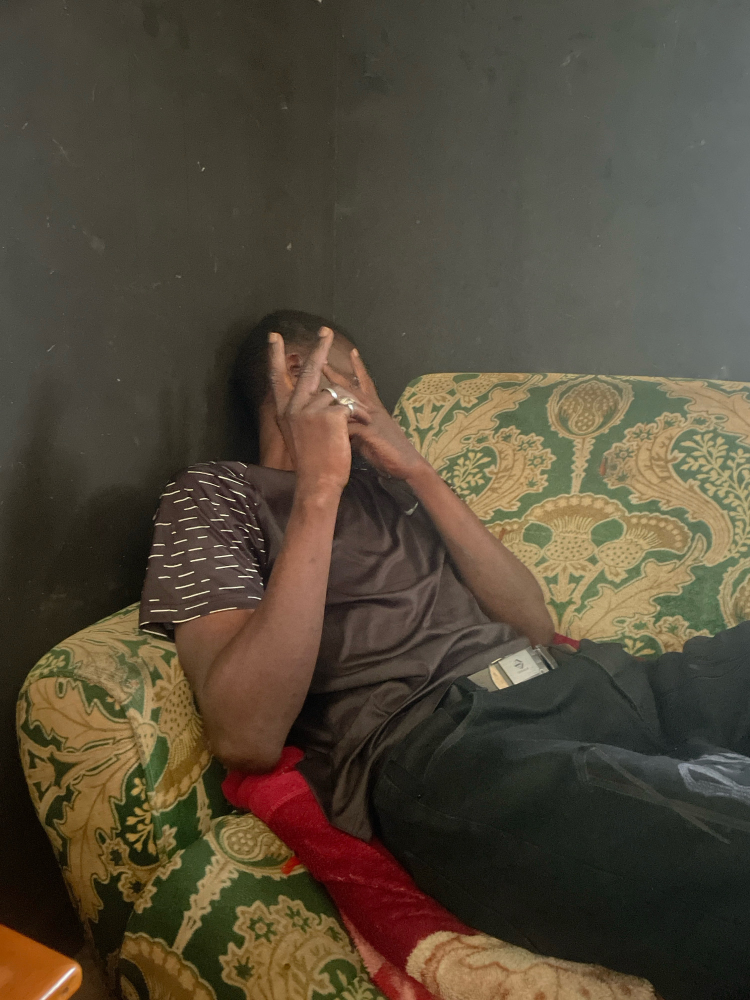 A young Black male is seated on a patterned couch. He is wearing a dark grey t-shirt with a white stripe pattern on the sleeves and black pants. His left arm is bent, and his hands are raised to his face, obscuring it. His right hand is near his face, with two fingers extended in a peace sign. He is wearing a silver ring on his right hand. A red piece of fabric is draped across his lap and over the armrest of the couch. The couch has a green and gold floral pattern. The background is a dark, textured wall. The scene appears to be indoors, possibly in a residential setting within the city of Jos, Nigeria.