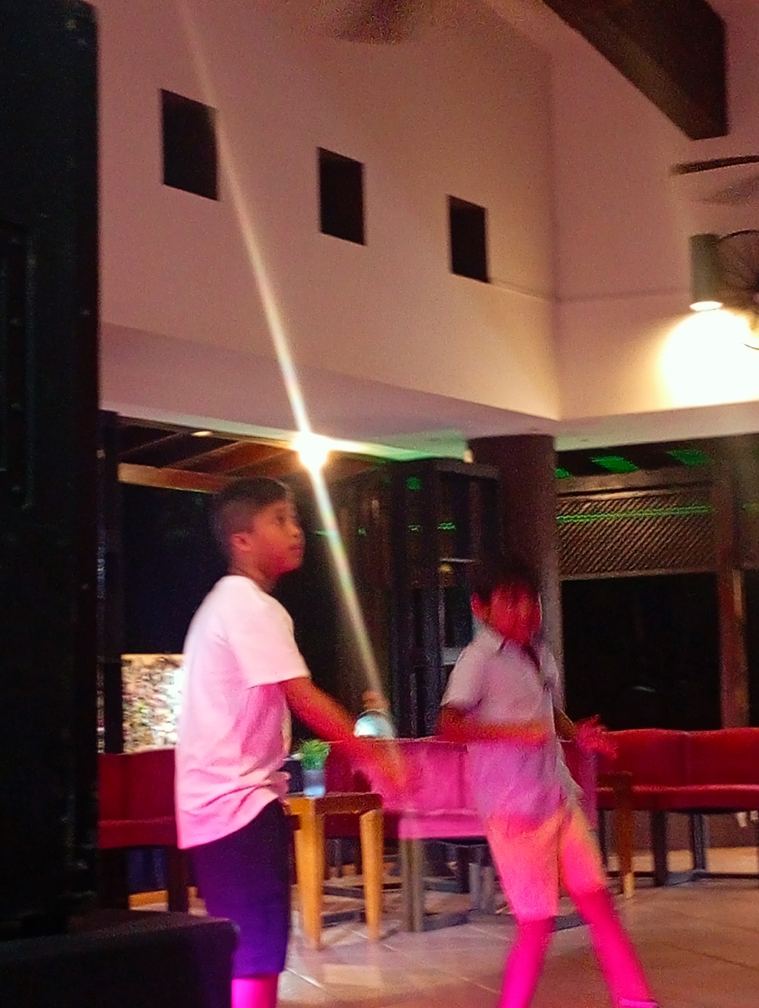 The image captures two young boys in an indoor setting, likely an event or performance space in Playa El Agua, Venezuela. The boy on the left wears a white t-shirt and dark shorts, looking to his right. The boy on the right wears a light-colored shirt and shorts, seemingly in motion. Red benches line the background. A dark structure is visible on the left side. The lighting in the space includes a ceiling light and potentially some color-changing lights that cast a pink hue on the boys and the floor. The architecture features square-shaped wall recesses. A ceiling fan can be seen in the upper right corner.