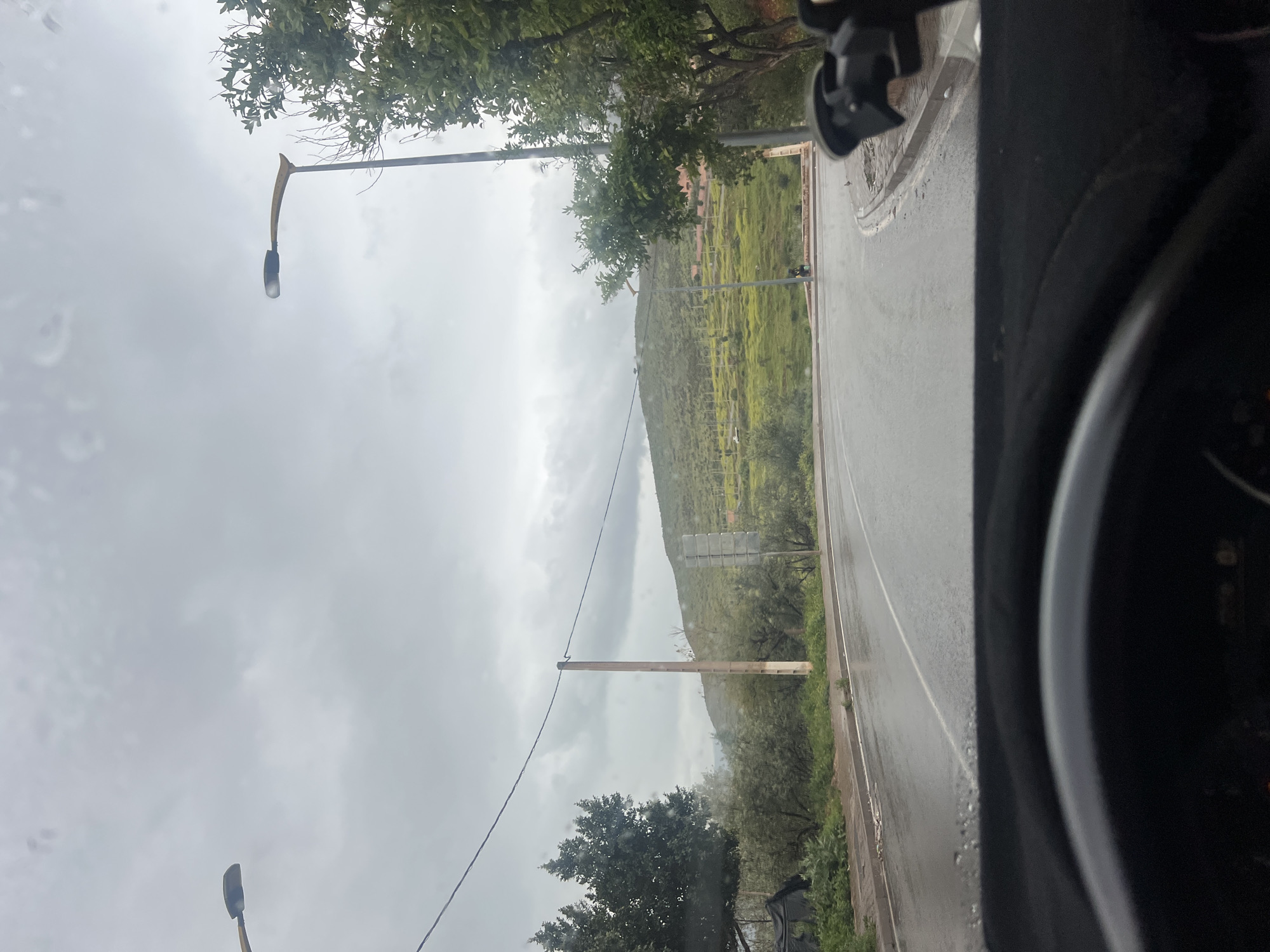 The image shows a view from inside a vehicle on a wet road near Ouaouizert, Morocco. A green hill rises in the background under a cloudy sky. There are utility poles alongside the road. The interior of the vehicle is visible on the right side of the image, including part of the dashboard and a phone mount. Street lights are visible in the image.