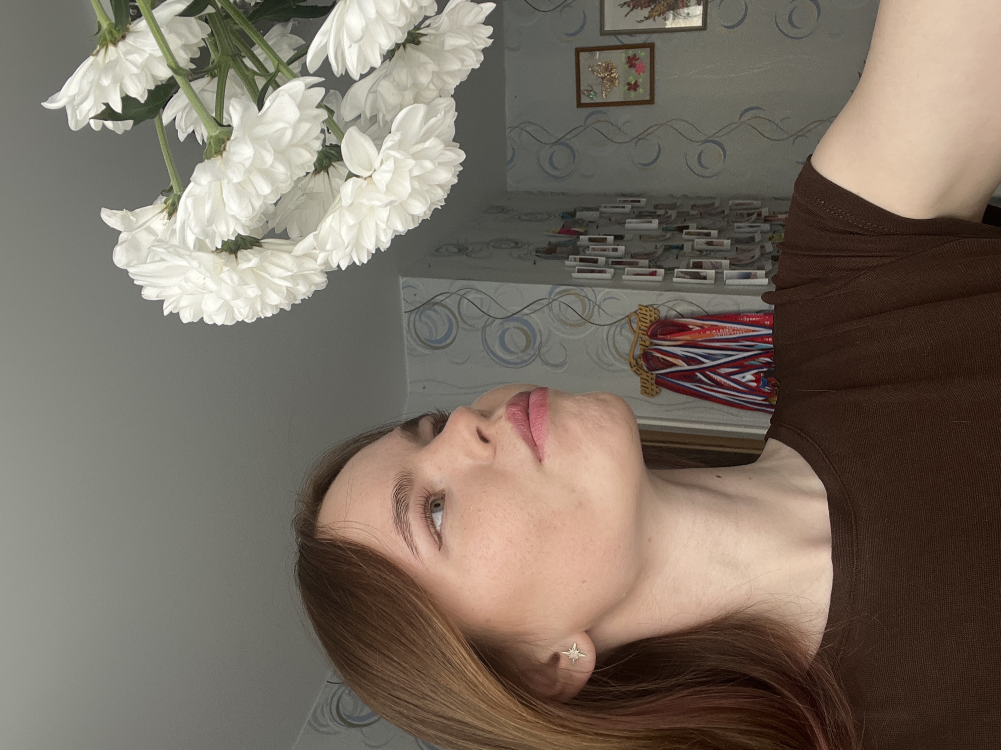 An indoor scene in Chaikovsky, Russia, displays a young Caucasian female from the upper torso, looking upwards and to the right. She has brown hair, light eyes, and freckles, wearing a star-shaped earring in her left earlobe and a plain brown short-sleeved top. Her lips are slightly pursed. A bouquet of white chrysanthemums with green stems is positioned above and to the right of her, within her line of sight. The background wall is covered in light-colored wallpaper featuring blue and grey circular patterns. Visible wall decorations include a vertical array of numerous small, rectangular items, colorful ribbons draped over a hook to the left, and a framed picture on the right depicting a gold butterfly and floral motifs. The ceiling is plain and light-colored.