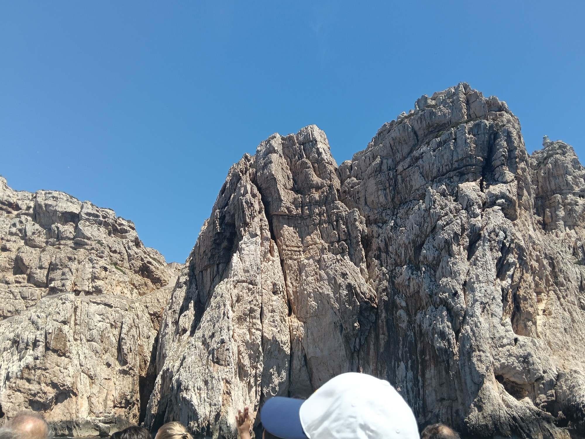 The image depicts a view of rugged, sun-drenched cliffs rising from the sea, under a clear blue sky. In the foreground, the heads and shoulders of several individuals are visible, suggesting they are on a boat. One person wears a white cap and another a blue cap. The rock formations are characterized by their textured surfaces, with visible cracks, crevices, and varied shades of brown and gray. The scale of the cliffs is imposing, emphasizing a natural, wild environment. The lighting suggests it is daytime, with strong sunlight casting shadows on the rocks. This scene is likely at Capo Caccia, a dramatic limestone promontory located northwest of Alghero, Sardinia, Italy, known for its spectacular cliffs and sea caves.