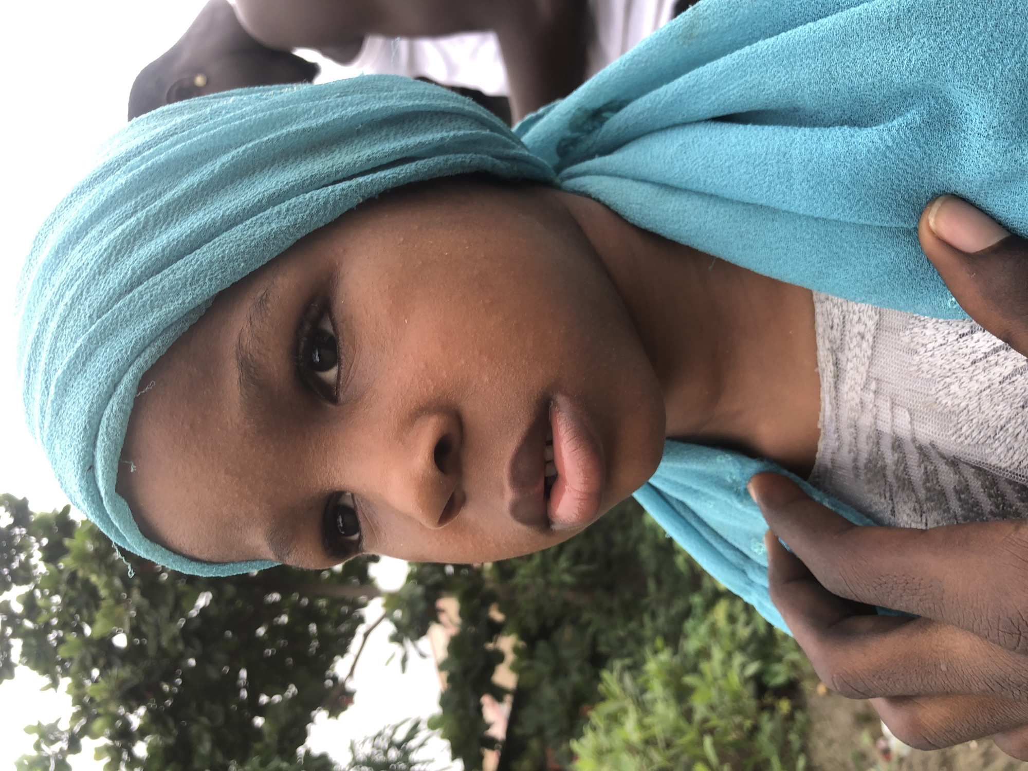 A close-up portrait captures a young girl with dark skin, wearing a teal headscarf and a grey textured shirt. Her gaze is directed off-camera to the left, her mouth slightly open. A hand with dark skin is visible in the lower left corner, gently touching the girl's chest. In the background, blurred green foliage and trees suggest an outdoor setting. The image appears to have been taken in Ado, Nigeria, based on the context.