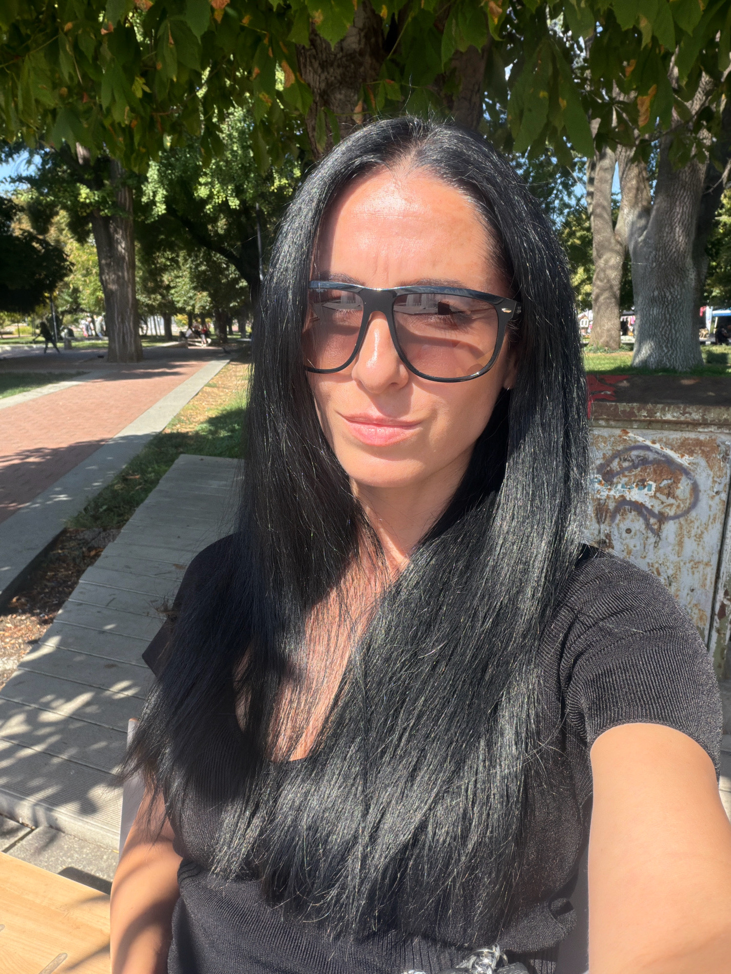 The image shows a woman taking a selfie outdoors in a park-like setting. She has long, dark hair and is wearing dark-rimmed sunglasses and a black top. Sunlight is casting shadows on her face and clothing. In the background, there are trees with green foliage, a paved walkway, and what appears to be a metal utility box with graffiti. The presence of mature trees, paved paths, and what might be park benches suggests a public park. Given the prompt's explicit request to identify the location as Pleven, Bulgaria, the scene is situated within a park in that city.