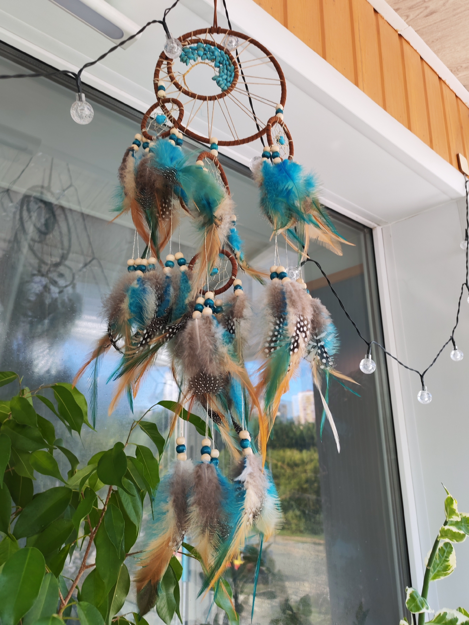 A dreamcatcher adorned with turquoise beads and feathers in shades of brown, blue, and tan hangs from a string of lights on the exterior of a building. The dreamcatcher is positioned near a window, with a green leafy plant in the foreground and a blurred background of trees and buildings. The building's exterior features wooden paneling and white trim. The scene is set in Izhevsk, Russia.
