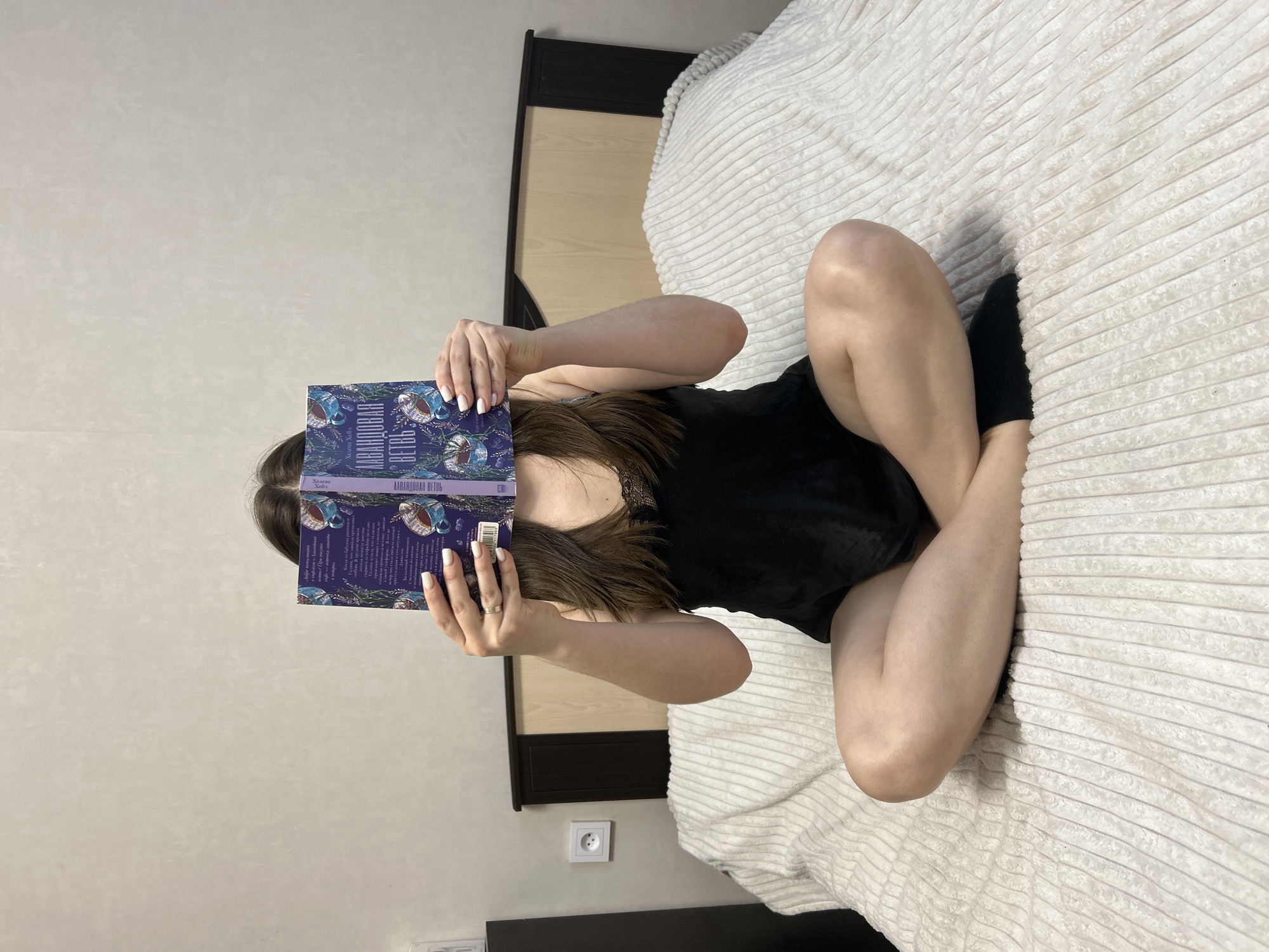 A person sits cross-legged on a bed, holding a book up to their face, obscuring it. The person is wearing a black top and dark socks. The book cover is purple and features a floral design with Cyrillic text, which reads "Лавандовая Ветвь" (Lavender Branch). The bed has a white, textured blanket. The scene appears to be indoors, in a bedroom or similar space, with a wall painted a light color. The location is in the city of Chaikovsky, Russia. A dark brown and light wood headboard is seen behind the bed. An electrical outlet is visible on the wall near the headboard.