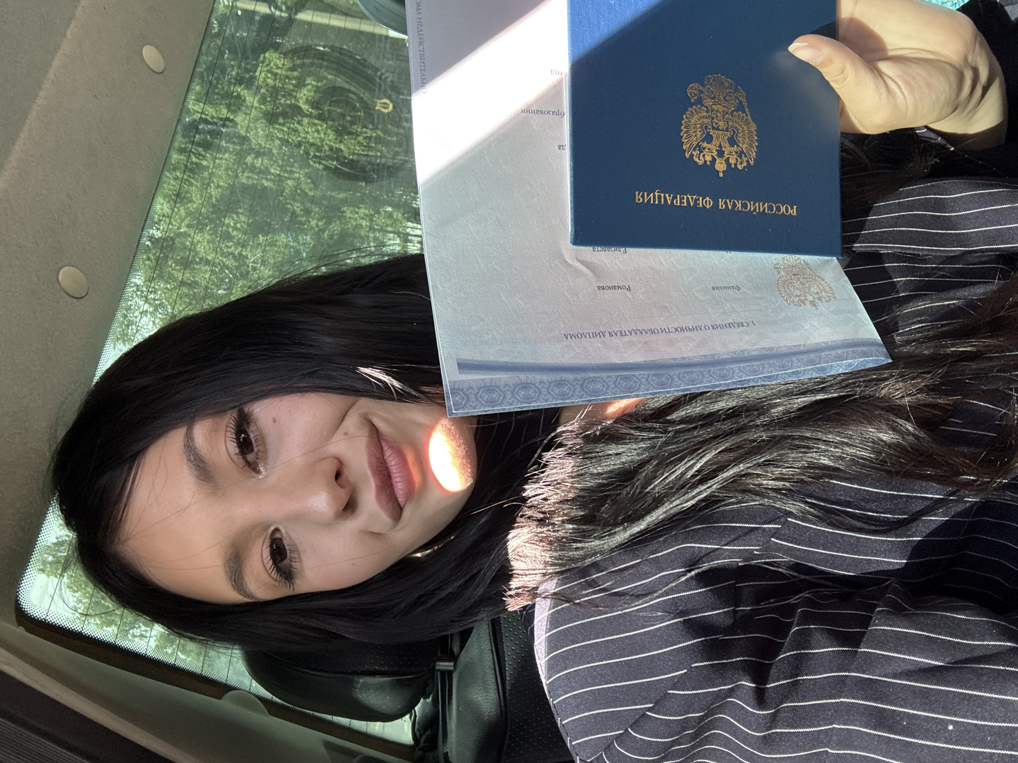 A young woman with long, dark hair and makeup is holding two documents. She is wearing a navy blue pinstripe jacket. One document is light blue and appears to be a diploma or certificate with Russian text. The other document is a dark blue booklet, also with Russian text and a gold emblem on the cover, which resembles a passport. She is sitting in the back seat of a car, evidenced by the headrest and the tinted window showing trees and sunlight outside. The overall scene suggests a celebratory or official occasion. The location is identified as Chaikovsky, Russia, based on the presence of a Russian passport.