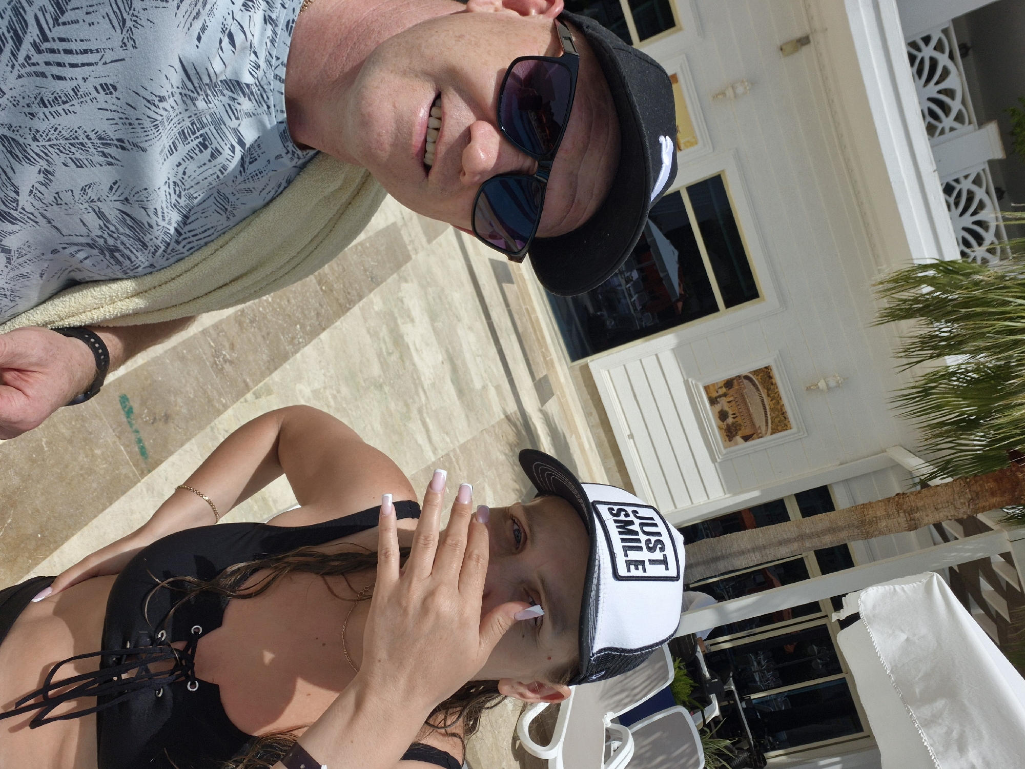 Two individuals are visible in the image, likely a male and a female, relaxing outdoors, possibly at a resort or hotel. The female is in the foreground wearing a black swimsuit, a gold chain, and a white and black trucker hat with the phrase "JUST SMILE" on it. She has manicured nails with a white tip and is partially covering her face with her hand. The male is in the background and is wearing sunglasses and a black cap. He is smiling. They are situated on a tiled surface in front of a white building with windows. There is a palm tree to the right. The location may be the city of Alanya, Turkey. There is furniture present, including chairs.