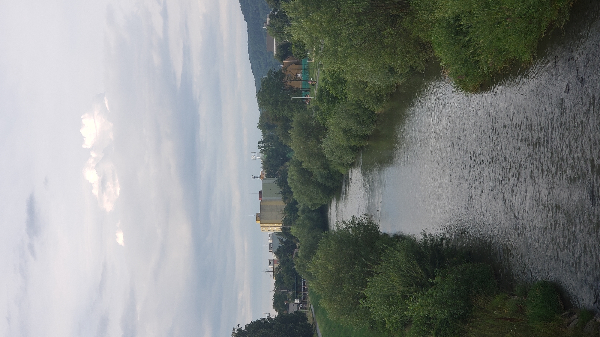 This is a vertical, medium-distance, eye-level shot of a river flowing through a city, likely Prešov, Slovakia. The river is the central focus, with its dark, rippling water reflecting the overcast sky. Lush green trees and bushes line both banks of the river. In the background, a city skyline is visible, featuring various buildings, including a prominent yellow and green multi-story structure, and several communication towers. Hills rise in the distance behind the city. The sky is filled with a mix of gray clouds and patches of bright white clouds, suggesting diffused sunlight. There are no people clearly visible in the image, though the hints suggest a path alongside the river where people might be present. The overall impression is of a peaceful, natural scene integrated within an urban environment.