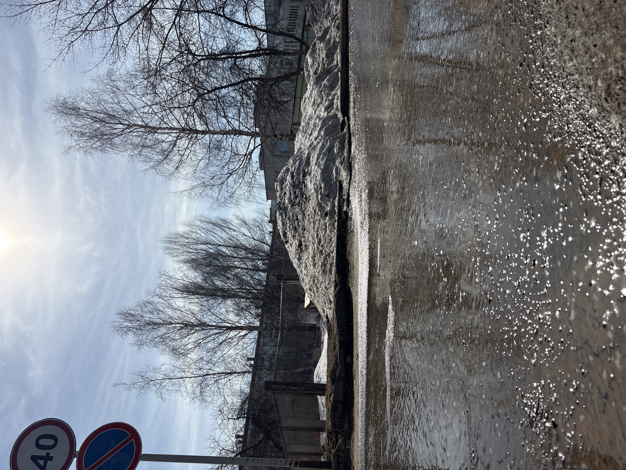 The image captures a street scene in Chaikovsky, Russia. A partially melted snowdrift lines the edge of the wet, reflective asphalt road. Bare trees and buildings are visible in the background, suggesting late winter or early spring. A "40" speed limit sign with a blue "no entry" symbol is in the foreground. The sky is partially cloudy, with the sun visible on the left.