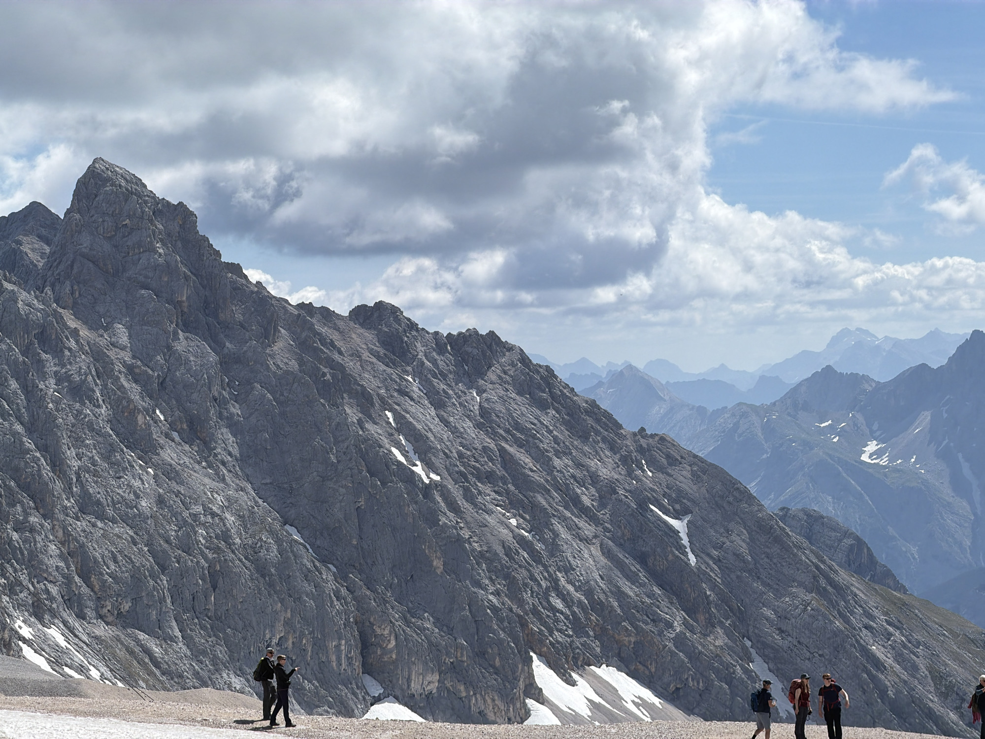The image depicts a dramatic mountain landscape with a steep, rocky slope dominating the foreground. Patches of snow cling to the mountain's surface, indicating a high altitude. In the distance, a series of layered mountain ranges recede into a hazy, blue atmosphere under a partly cloudy sky. Several individuals are visible on a gravelly plateau at the base of the mountain, appearing to be hikers. They are dressed in outdoor clothing and some are carrying backpacks. The scene is likely located in the Zugspitze region near Garmisch-Partenkirchen, Germany, given the characteristic alpine terrain and the typical activities of visitors to this area.