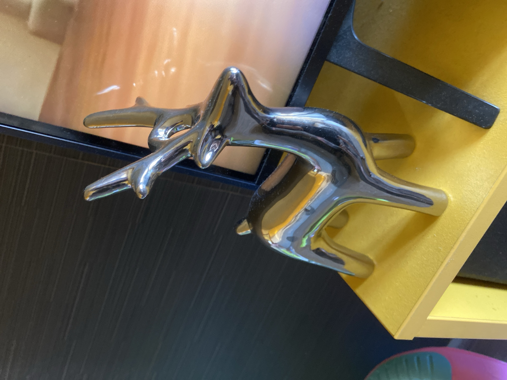 This media file depicts a close-up, angled view of a silver, stylized sculpture resembling two intertwined deer or stags. The sculpture has a sleek, polished finish that reflects the surrounding environment. It is positioned on a bright yellow shelf or surface, which appears to be part of a display or furniture. Behind the sculpture, a dark, textured surface is visible, possibly a wall or a piece of cabinetry. Further in the background, a portion of an object with a soft, peachy-orange color and a gentle gradient is visible, likely a lamp shade or a decorative element. The lighting suggests an indoor setting, possibly during daylight hours, as indicated by the reflections and the overall brightness. There are no visible people, text, or specific time of day or weather cues beyond the indoor lighting. The scene is focused on the metallic art piece and its immediate surroundings. The style of the sculpture suggests it could be a decorative item for a home or gallery.