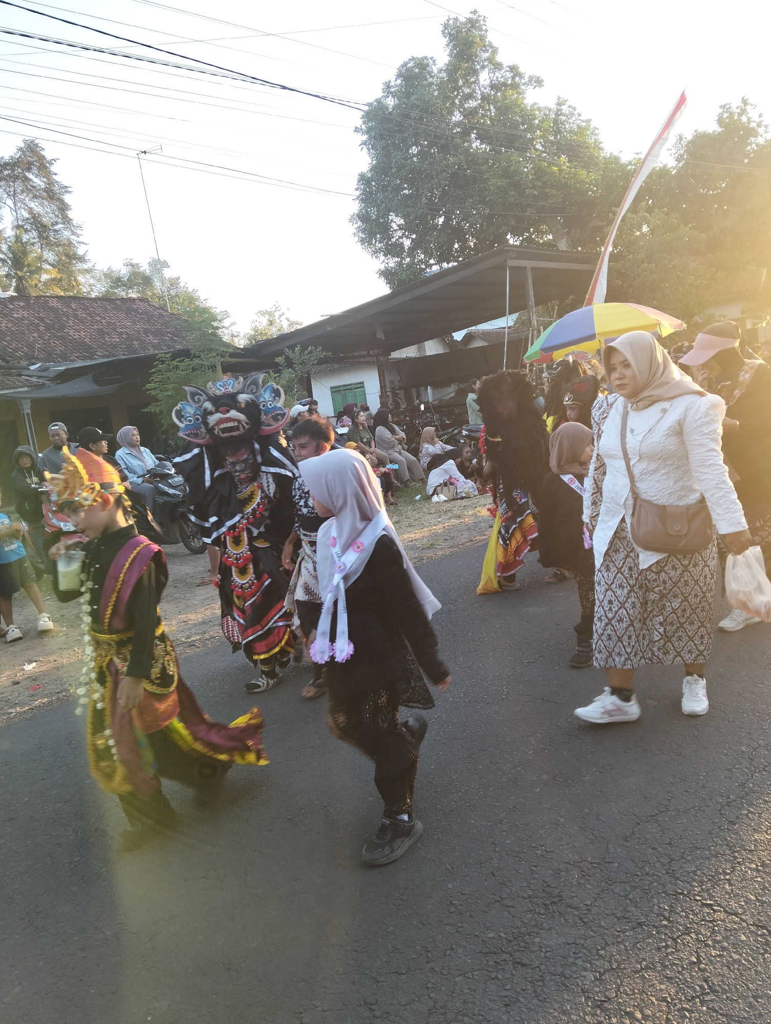 This is a candid, eye-level, full-shot photograph taken outdoors in what appears to be the city of Sumbersewu, Indonesia, during daylight hours. The scene captures a street procession, likely part of a cultural event or festival. The foreground features several individuals dressed in traditional attire. To the left, a young person in a decorative headdress and elaborate, colorful garment is walking, holding a drink. To the center, another individual is wearing a dark costume with bright red and yellow accents, topped with a large, fierce-looking mask resembling a mythical creature, possibly a barong. Their legs are adorned with striped leggings and traditional shoes. To the right, a woman wearing a light-colored hijab and a patterned skirt is walking, carrying a light brown purse. Behind these figures, other participants and onlookers are visible, some in traditional clothing and others in casual wear. There are also motorcycles and makeshift shelters along the side of the road. The background shows houses with tiled roofs and lush green trees, typical of a rural or semi-urban Indonesian setting. The sun is low in the sky, casting a warm glow and creating lens flare in the upper right portion of the image. The overall atmosphere suggests a lively community celebration.