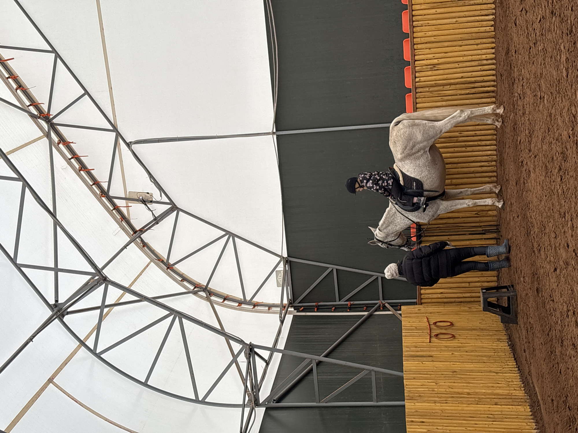 The image shows an indoor equestrian arena in Русский Вожой, Russia. A rider wearing a black helmet and a patterned shirt is seated on a light gray horse. A person in a black jacket and white hat stands near the horse. The arena has a sand floor and wood panel walls, with orange markers attached to them. The roof of the arena is a white and gray tent-like structure with a visible metal framework.