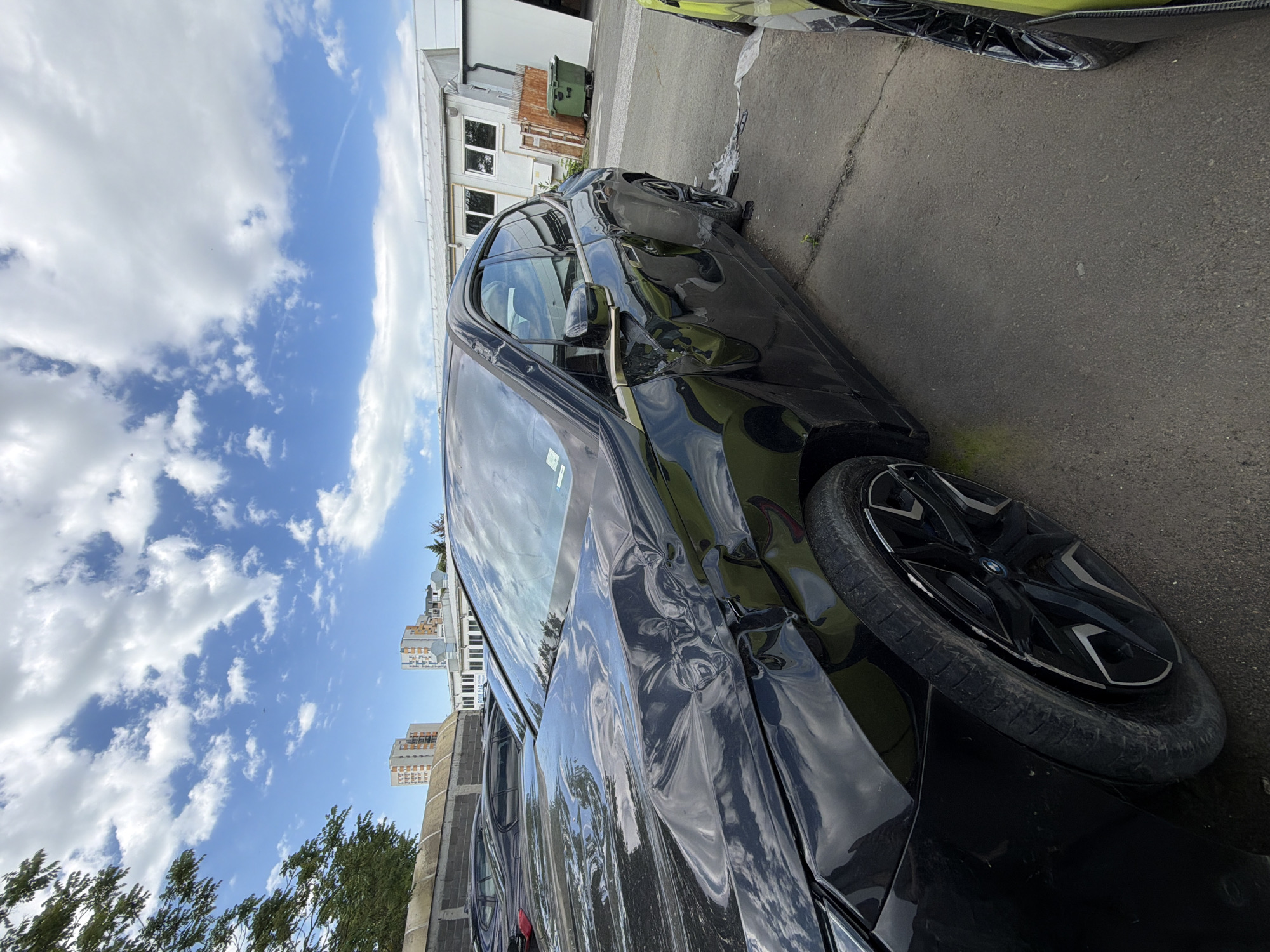 The image shows a severely damaged black car on an asphalt surface under a partly cloudy sky. The car's front end and driver's side have extensive crumpling and deformation. The car appears to be next to a building and another vehicle with some type of green wrap. In the background are buildings and trees. There is no visible evidence to verify the location in Burgas, Bulgaria. No people are visible.