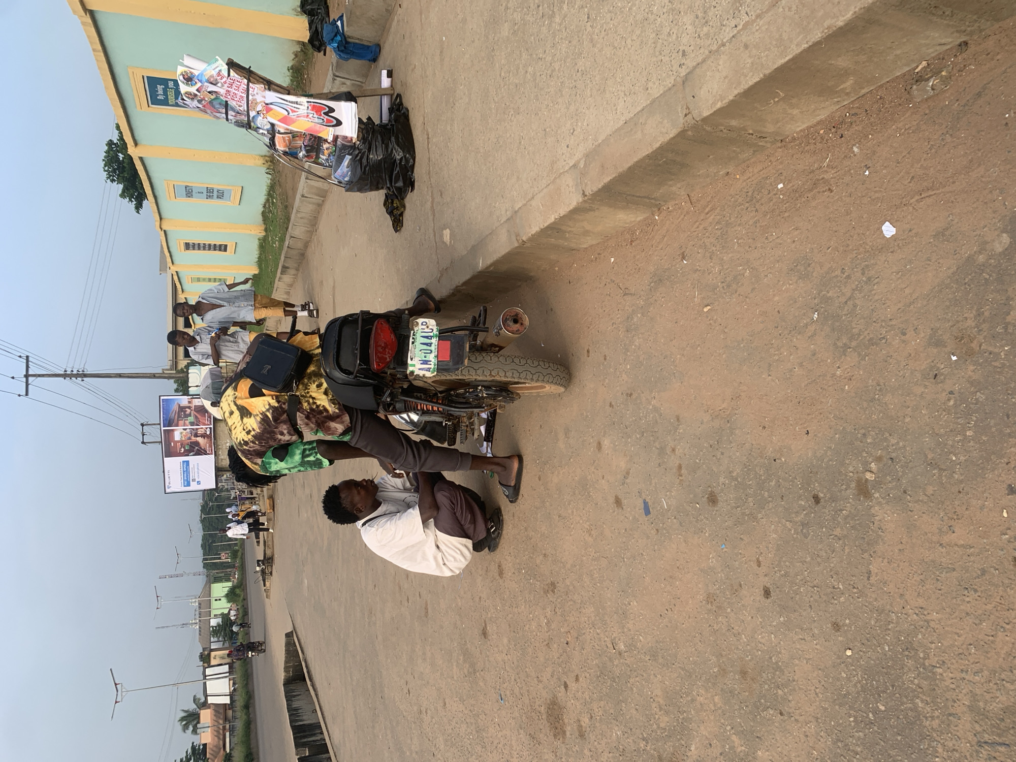 In Shagamu, Nigeria, on a paved roadside, two men are interacting near a motorcycle. One man, wearing a white t-shirt and dark pants, squats down to face the motorcycle's rear wheel, holding a thin object in his mouth. The second man, dressed in a colorful patterned shirt and dark pants, stands beside the motorcycle, looking down at it. The motorcycle has a license plate reading "AN-0444-UP". To the right of the motorcycle, a rack displays various items for sale, including magazines and posters. Two young boys walk along the sidewalk in the background, passing a light green wall with yellow trim adorned with posters. The environment appears to be an urban setting with utility poles and streetlights visible.