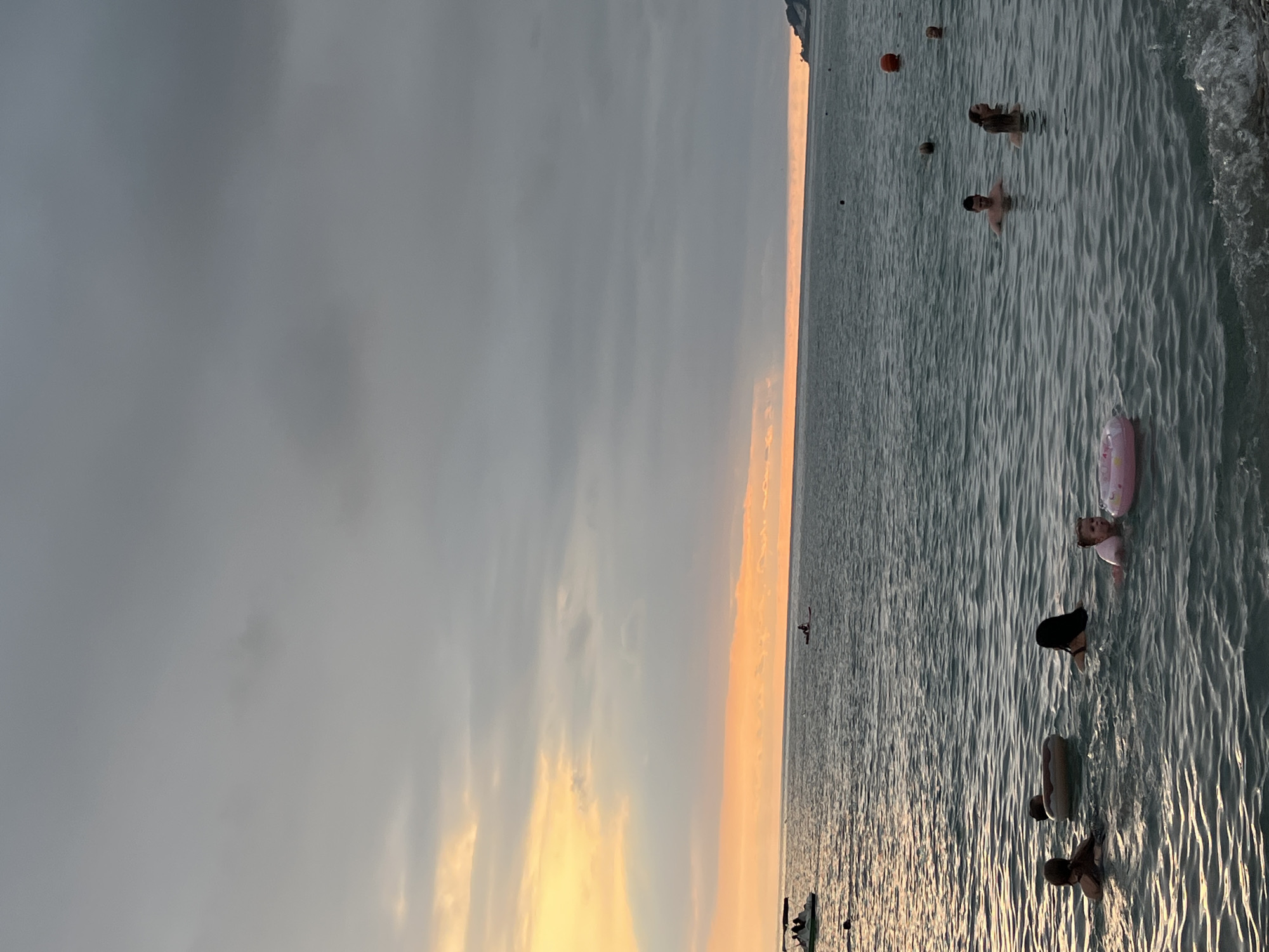 The image depicts a coastal scene, likely the city of Sochi, Russia, during what appears to be sunset. The sky is filled with dramatic clouds, with a band of orange and yellow light near the horizon, suggesting the sun is setting. The sea is calm, with gentle waves lapping at the shore in the foreground.
Several people are visible in the water. In the foreground, a child wearing a pink inflatable ring is being supported by an adult. To their left, another person is swimming, and further left, another individual is partially submerged, with an inflatable ring nearby. In the mid-ground, a group of people are swimming and interacting. There are also a few scattered buoys or markers in the water.
Further out in the sea, a small boat with people on board is visible on the left, and on the right, a person is on a paddleboard or kayak. In the distance, a coastline with some vegetation and structures is faintly visible, and the sea meets the horizon under a cloudy sky. The overall impression is one of a relaxed evening at the beach.