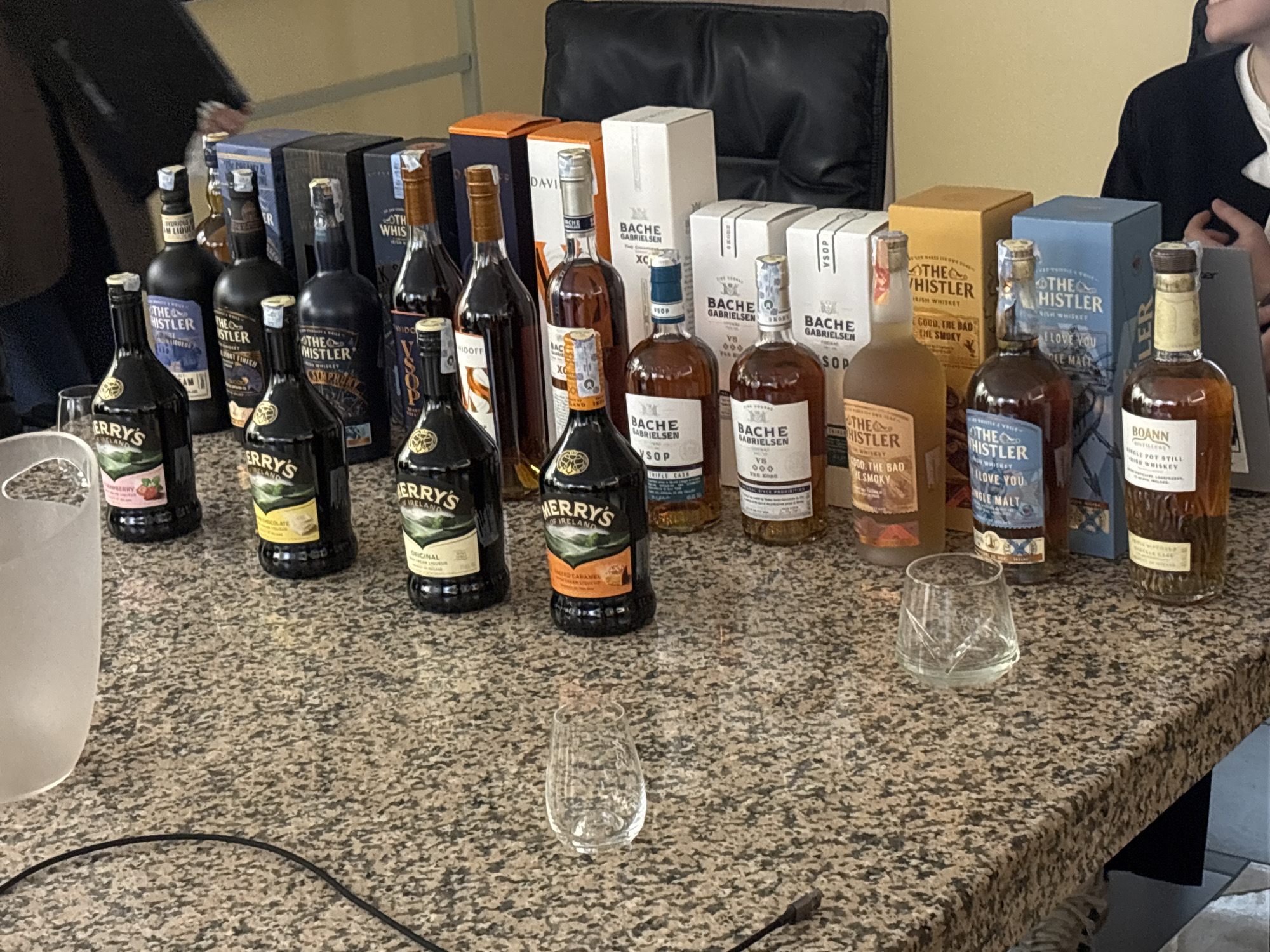 The image shows a collection of liquor bottles arranged on a countertop with a speckled, granite-like surface. There are several bottles of "Kerry's" liqueur and "The Whistler" Irish whiskey, as well as "Bache Gabrielsen" cognac. Boxes corresponding to some of the bottles are placed behind them. Three empty glasses sit on the counter. The setting appears to be indoors, possibly in a tasting room or private residence. A person in a brown jacket and dark pants stands to the left, while another person in a black jacket stands to the right. A black leather chair is behind the bottles. The location is not evident from the image.