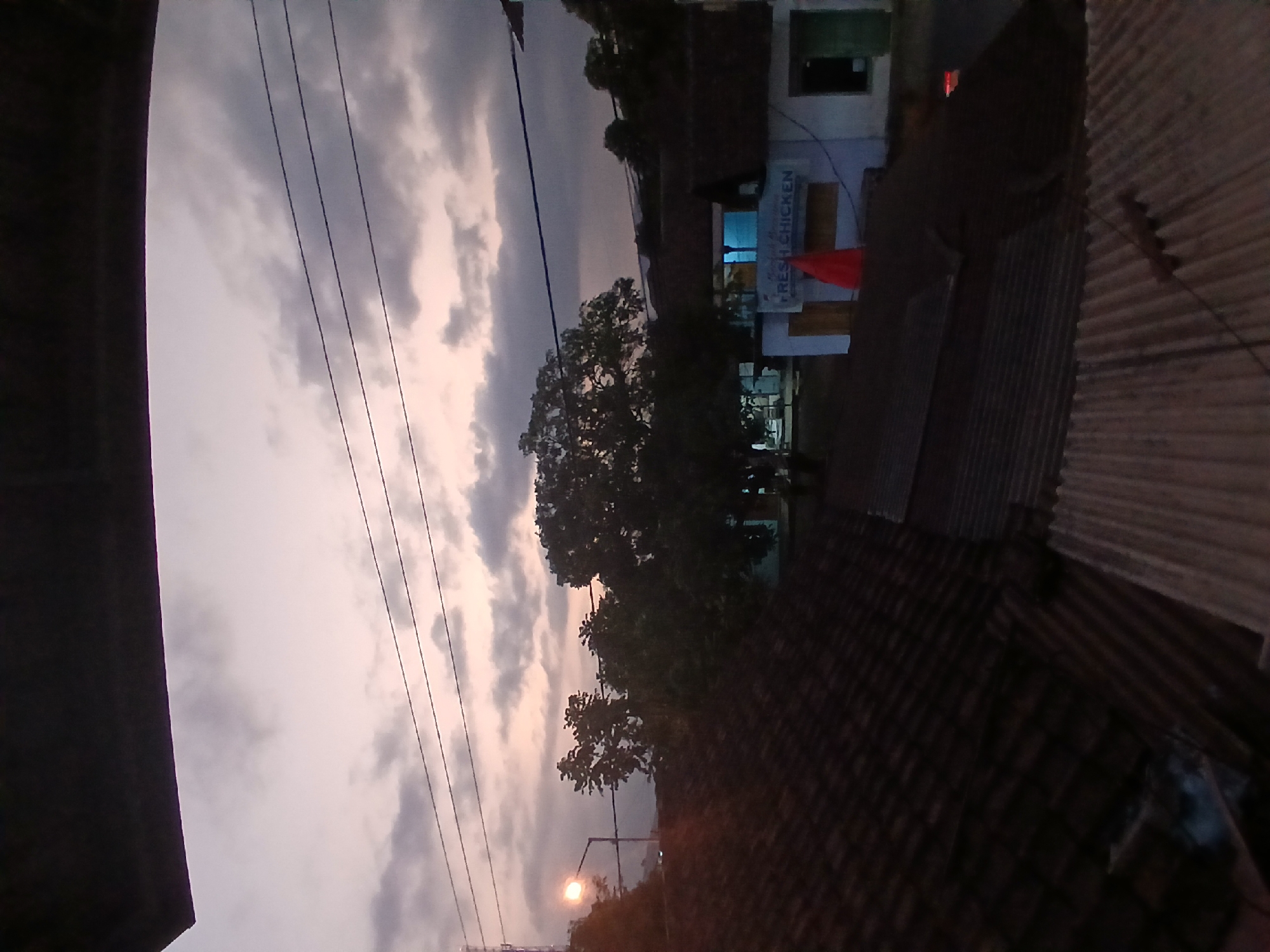 The image depicts a street scene at dusk in Kademangan Kulon, Indonesia. A street lamp illuminates the area, casting a glow on a large tree. In the background, a building with a sign that reads "FRESH CHICKEN" is visible. A red flag is placed in front of the building. The foreground is dominated by rooftops of houses. The sky is overcast with clouds. The overall atmosphere is dim and tranquil.