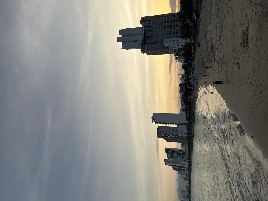 The image depicts a wide shot of a coastal urban landscape at twilight in Nha Trang, Vietnam. The sky transitions from a light blue-grey at the top to a warm gradient of yellow and orange near the horizon. A calm body of water, likely the South China Sea, occupies the lower left and center, exhibiting gentle waves breaking onto a sandy beach.
The coastline is lined with a dense array of modern high-rise buildings, including multiple skyscrapers and hotels, extending into the distance. A prominent multi-story building with a lighter upper section and a darker, textured lower section stands on the far right. Distant mountains are faintly visible behind the city structures on the left horizon.
On the sandy beach, approximately 10-15 individuals are observed. Several people are walking on the sand, while others are in the shallow water, wading or swimming. One person in dark attire walks on the mid-foreground sand towards the viewer. Along the back edge of the beach, near the base of the city buildings, a line of tents, umbrellas, or temporary shelters is visible. On the far right of the beach, several dark, covered objects, resembling small boats or fishing vessels, rest on the sand.