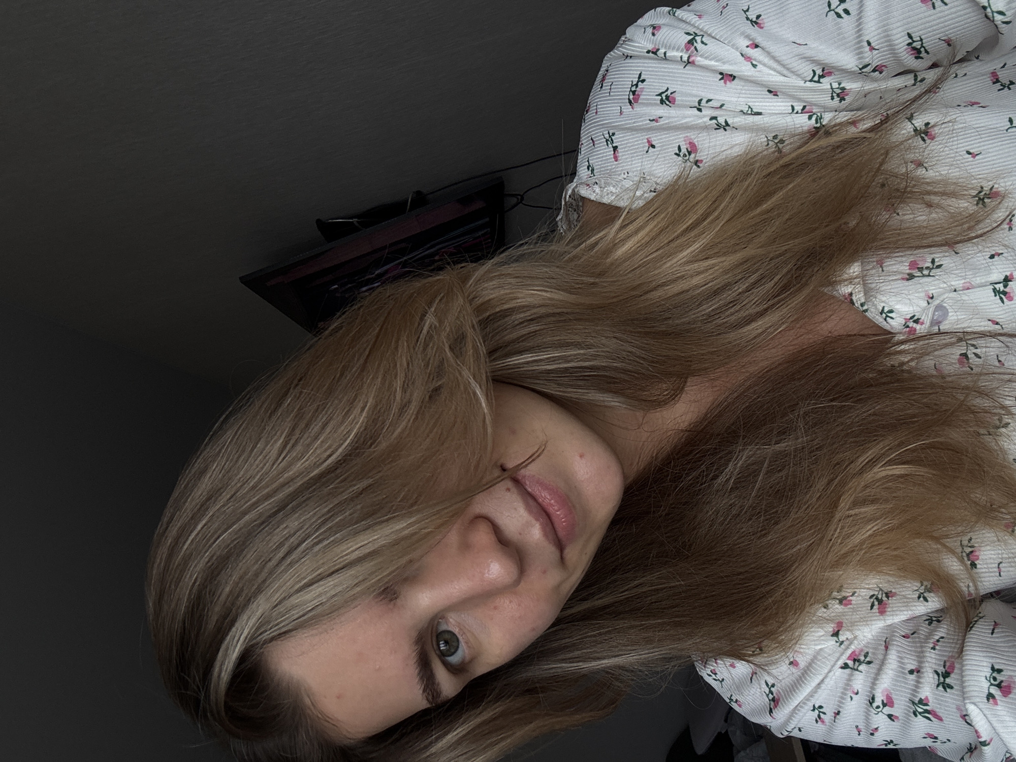 A person with long, blonde hair and a floral-patterned white shirt is visible. The person is indoors, in a room with a dark-colored wall and a television. The location appears to be in the city of Unknown, Unknown.