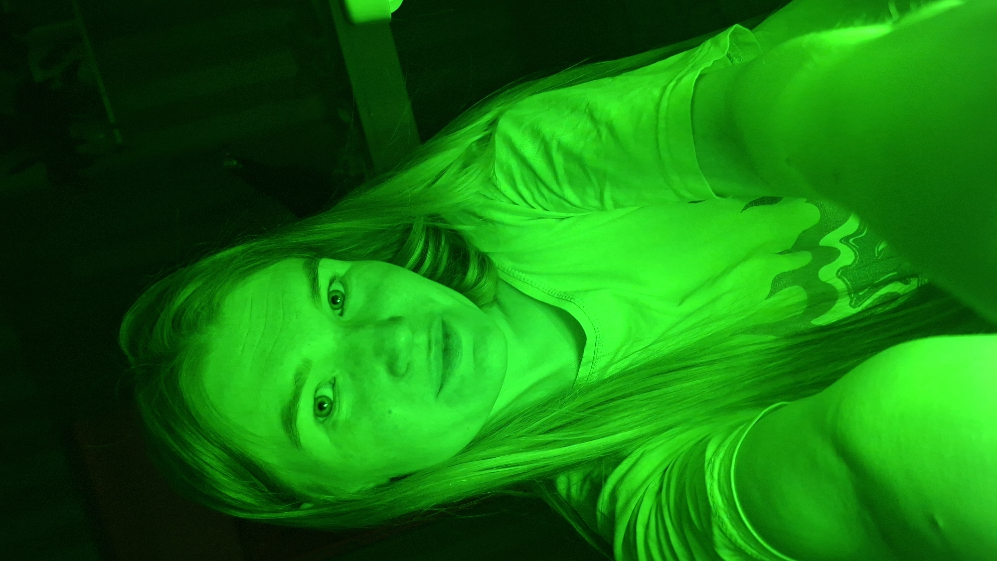 The image displays a young woman illuminated by an intense, vibrant green light, giving the entire scene a monochromatic and stylized appearance. She is positioned diagonally, lying down with her head towards the lower left of the frame. Her long, light-colored hair is spread out, reflecting the green light prominently. She looks directly at the viewer with an open-eyed, neutral expression. She is wearing a t-shirt, which appears bright green, and a subtle, wavy pattern can be faintly discerned on the fabric in the lower right portion of the image. The background is dark and largely indistinct, suggesting an indoor or dimly lit setting. No specific objects or structures are clearly visible in the background apart from vague, dark shapes. There is no visible text. The image was captured in Большой Букор, Russia.