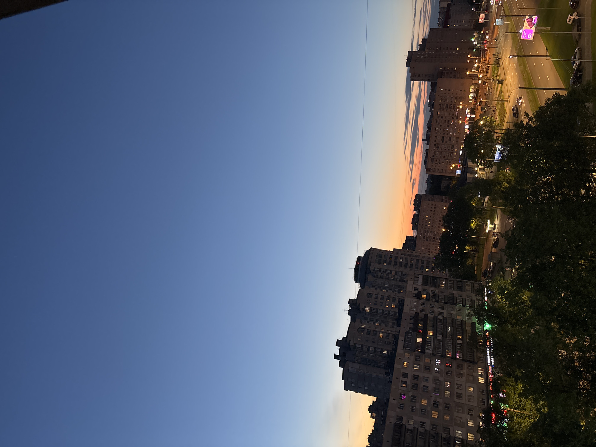 The image depicts a cityscape at dusk. In the lower-right quadrant, there are densely packed apartment buildings, with numerous windows illuminated. A road lined with streetlights stretches across the right side, and vehicle traffic can be seen. Trees obscure parts of the lower-right corner. The sky transitions from a gradient of orange and yellow near the buildings to a deep blue in the upper-left, indicating the time of day. The scene appears to be of a residential area. Based on the cityscape and architecture, it is likely to be in a city like Vsevolozhsk, Russia. No people are clearly visible in the scene.
