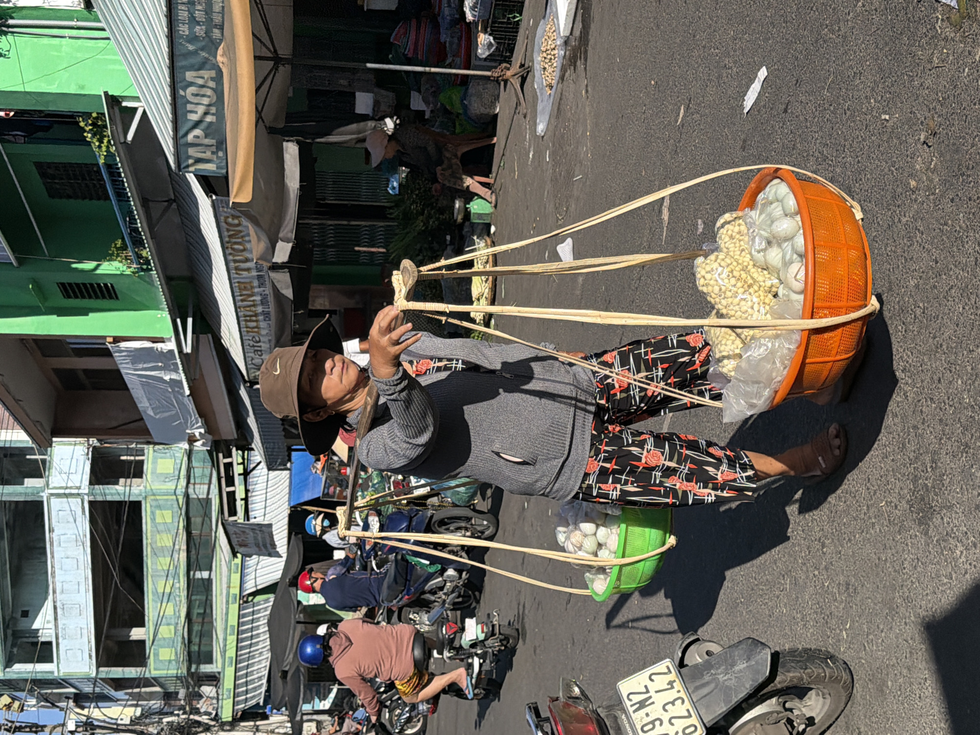 A Vietnamese woman is walking down a street in Nha Trang, Vietnam, carrying a yoke across her shoulders with two large baskets of produce. The woman is wearing a wide-brimmed hat, a gray cardigan, and patterned pants. The baskets contain what appear to be eggs and some other food items. The street scene includes several motorcycles, buildings with awnings, and other vendors in the background. The ground is paved, and sunlight casts shadows. The overall impression is of a busy market street.