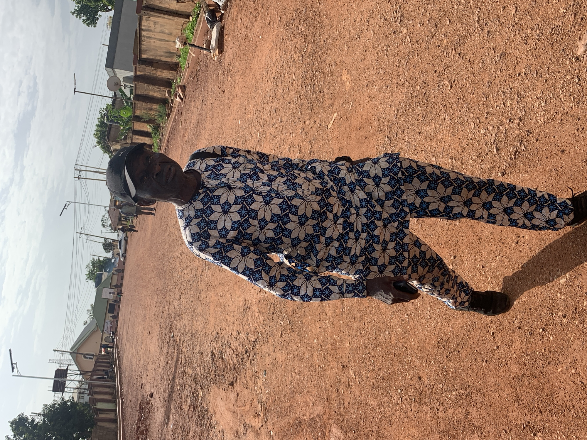 In the city of Gura Top, Nigeria, an elderly man is captured in a medium shot, standing on a reddish-brown dirt road. He is wearing a patterned traditional outfit in white, blue, and black, with a black baseball cap. His left hand is holding a dark-colored object, possibly a mobile phone. The man is looking directly at the camera with a serious expression. The background depicts a street with buildings, power lines, and some trees under a partly cloudy sky. The lighting suggests it is daytime.