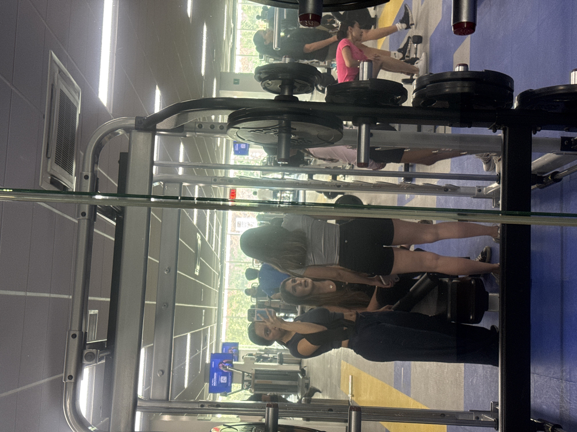 The image depicts a gym setting, presumably in Puebla, Mexico. Through a glass partition that causes reflections, several individuals are visible. In the foreground, a woman in dark attire is taking a picture with her phone, likely a selfie. Behind her, another woman wearing a gray shirt and black shorts appears to be interacting or posing with the selfie-taker. In the background, there are other individuals using gym equipment, including one wearing a pink shirt and doing leg exercises on a machine. The gym has various weightlifting machines with stacked weight plates. The floor has a combination of blue and yellow sections, indicating different areas or activity zones within the gym. The ceiling features rectangular light fixtures and what appears to be an air conditioning unit. There are windows letting in natural light, partially obscured by tree foliage outside.