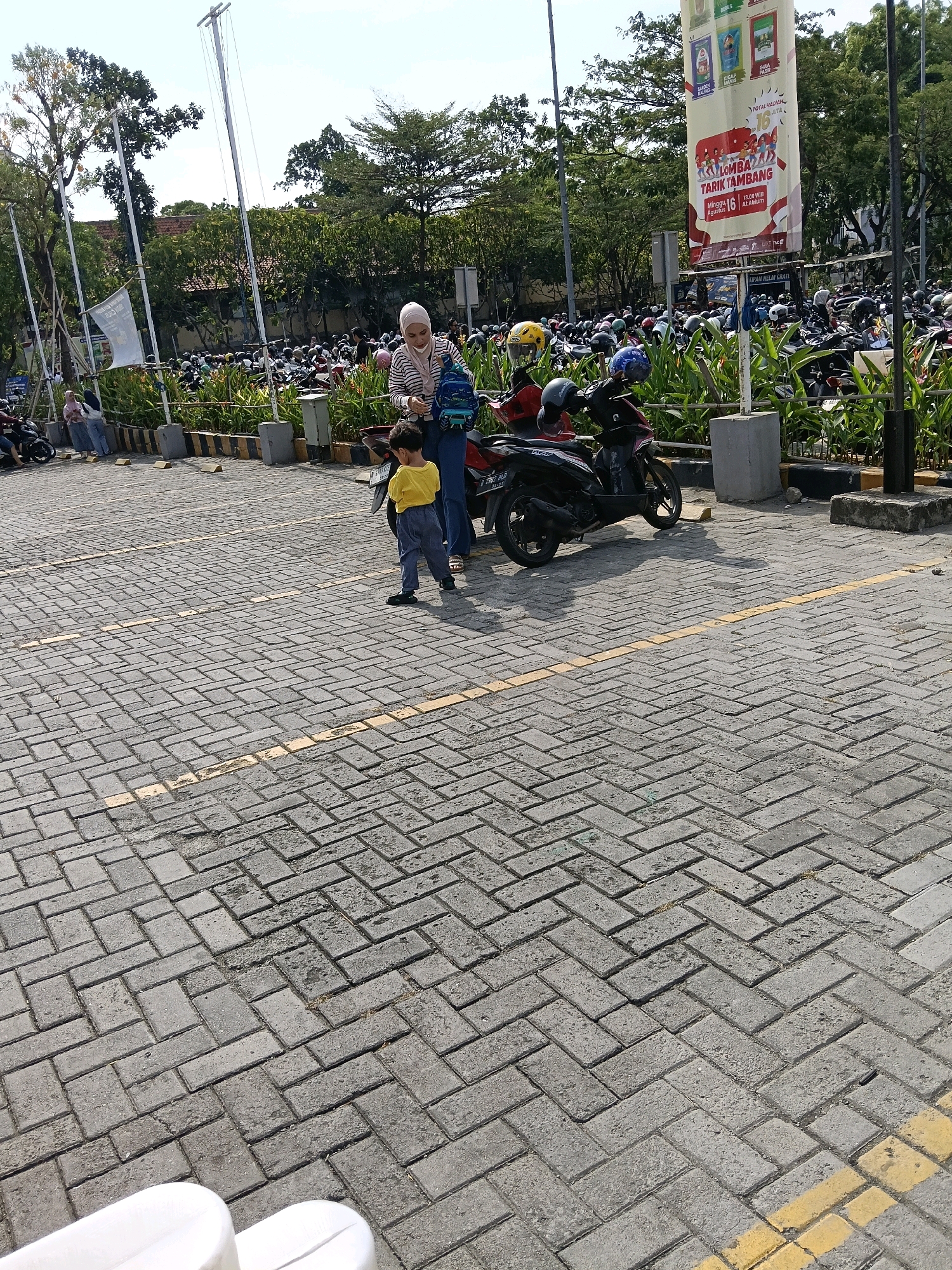The image depicts a busy outdoor parking lot, likely in Tangerang, Indonesia, based on the large banner visible in the background. The banner is predominantly white and red, featuring text and images advertising a "TARIK TAMBANG" (tug-of-war) event scheduled for Sunday, August 16th, at 13:00 WIB at "At Alun". This suggests the event is a community or promotional activity.
In the foreground, a woman wearing a hijab and a striped long-sleeved shirt, along with a young boy in a yellow t-shirt and gray pants, are standing near a parked motorcycle. The woman appears to be adjusting something on her backpack, while the boy is looking away from the camera. The motorcycle is dark-colored with a red accent on the seat. The ground is paved with interlocking gray bricks, marked with yellow lines, indicating parking stalls.
Behind them, a vast number of motorcycles are parked, filling the lot. The scene is bordered by lush green foliage and trees. In the distance, more trees and some buildings are visible, as well as several tall poles with wires, possibly for lighting or power. The sky is bright and clear, suggesting a sunny day. The overall atmosphere is one of a public gathering or a commercial area with significant motorcycle traffic.