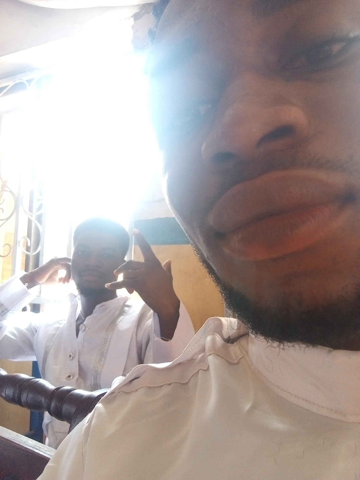 A close-up, low-angle selfie captures two dark-skinned adult males indoors, heavily backlit by a prominent window. The foreground subject, positioned to the right and lower frame, is extremely close to the camera, with only the lower portion of his face visible, including his full lips, nose, and chin with short facial hair. He wears a white collared garment, possibly a traditional tunic. A dark brown, carved wooden railing occupies the lower left corner.
In the background, positioned to the left and center, the second male is visible from the chest up, silhouetted against a bright, overexposed window with ornate, dark metal security bars or a decorative grill. He also wears a white long-sleeved shirt or tunic, featuring silver-colored trim or embroidery on the placket and what appears to be a metallic necklace. His right hand is raised, performing a "rock on" or "devil horns" gesture with the index and pinky fingers extended. His facial features are obscured by the intense backlighting, but he appears to be looking towards the camera. The wall behind him and to his right is painted light blue, transitioning to a light tan or brown lower down. The extreme brightness from the window creates significant lens flare and haze across the upper and central portions of the image.