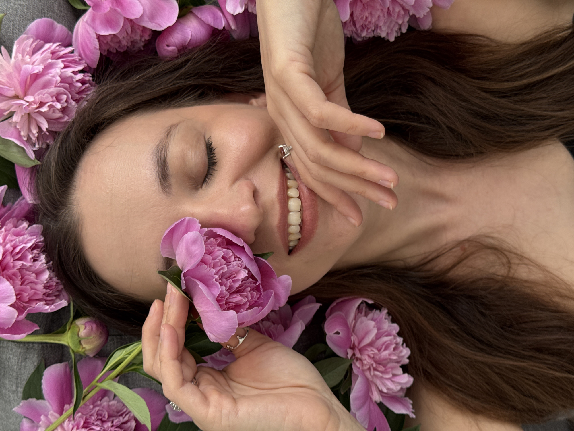 A woman with long brown hair is lying on a gray surface surrounded by pink peonies. Her eyes are closed, and she is smiling slightly, with a peony held up to her face with her left hand, on which she is wearing a silver ring. Her right hand is raised near her mouth. The scene likely takes place indoors. There is no evidence to support it being located in Chaikovsky, Russia.