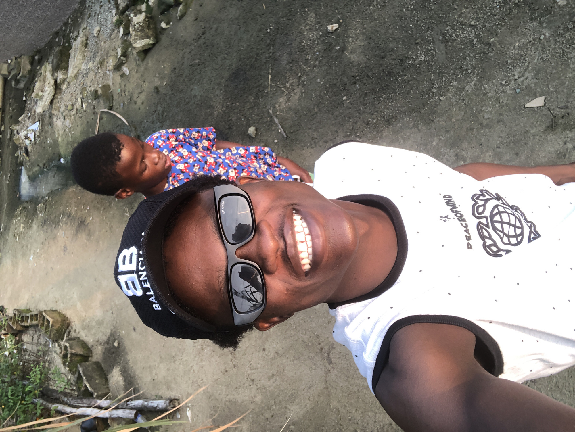 The image is a selfie taken by a young Black male, wearing a black baseball cap with "BALENCIAGA" printed on it in white letters, dark sunglasses with "off white" written on the lenses, and a white sleeveless shirt with small black dots and a graphic of a globe with text "PEACE OF MIND". He is smiling broadly and looking at the camera. Behind him, to the right, a young Black child, likely a boy, is standing and looking downwards. The child is wearing a blue shirt with a red floral pattern. The background appears to be an unpaved path or street with debris, rocks, and some vegetation. The setting suggests an outdoor urban environment, possibly in a less developed area. The lighting indicates it is daytime, possibly late afternoon due to the shadows. The overall impression is informal and candid.