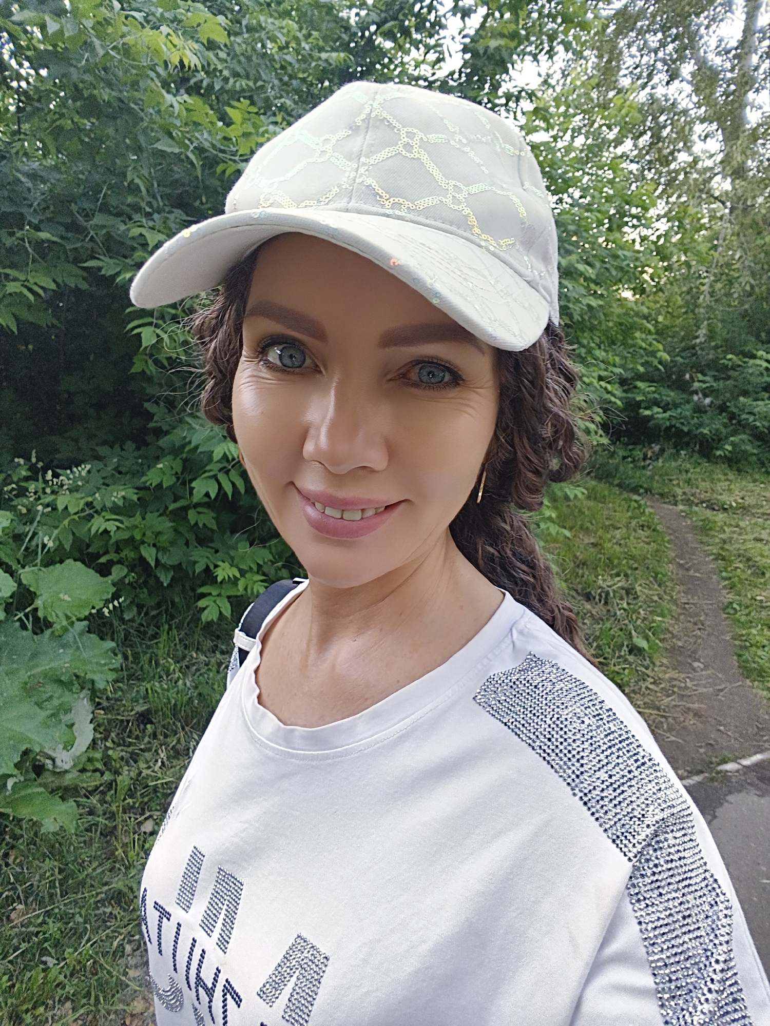 A woman is pictured outdoors, in a medium shot, wearing a white baseball cap with a geometric sequined pattern and a white t-shirt with sequined detailing on the shoulders and chest. She has curly brown hair, blue eyes, and is smiling. She is wearing a gold hoop earring. The background consists of lush green foliage and a dirt path. The lighting suggests it is daytime, with soft natural light illuminating the scene. The setting appears to be a park or a wooded area.